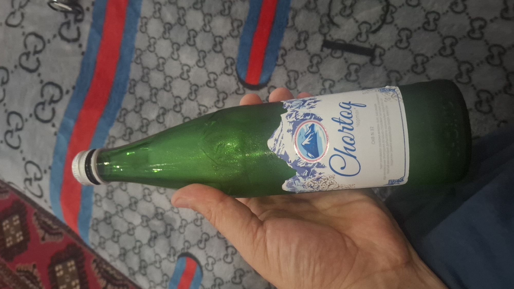 A person's hand holds a green glass bottle of Chartoq brand mineral water. The bottle has a white label with a mountain graphic and blue text. The background shows a fabric with a repeating pattern of the Gucci logo, and a striped blanket in red, blue, and white. The image is a close-up of the bottle and hand, suggesting it was taken indoors. The bottle is likely located in Yangi Abad, Uzbekistan, as this is the origin of Chartoq mineral water.