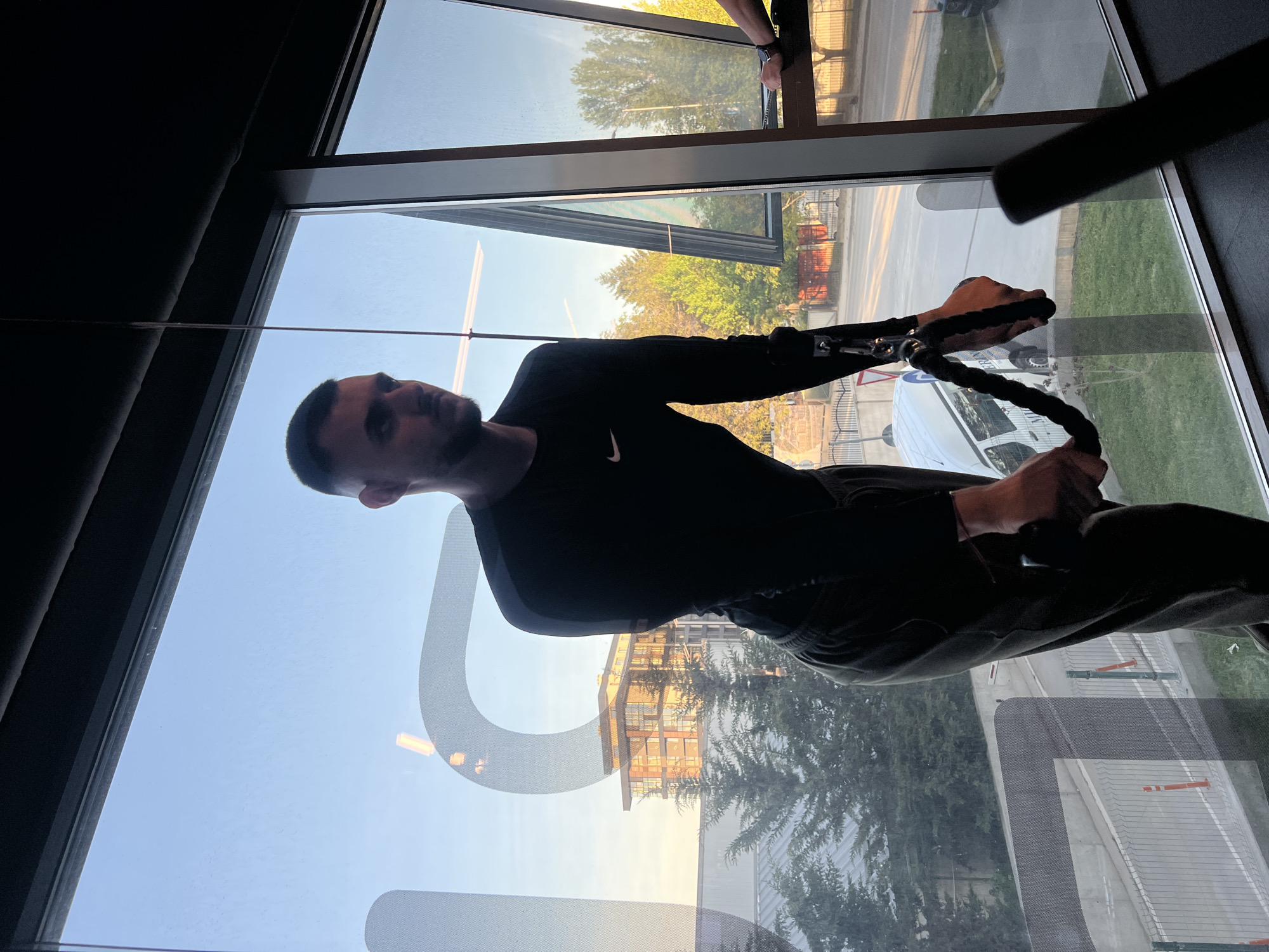 The image, taken in Plovdiv, Bulgaria, captures a man exercising indoors next to a large window overlooking an outdoor scene. The man, appearing to be in his 20s or 30s with short dark hair and a beard, is wearing a black long-sleeved shirt with a white Nike logo on the left chest and dark athletic pants. He is actively engaged in a workout, gripping a black rope attachment from a cable machine with both hands, suggesting a pulling or pushing exercise. His expression is one of focus and concentration.
Through the large window, a clear outdoor setting is visible. The sky is a light blue, indicative of good weather. Distant buildings and lush green trees are illuminated by the warm, golden light of what appears to be late afternoon or early evening. Below the trees, a paved road or pathway with grassy verges and some orange traffic markers can be seen. In the upper right section of the window, the arm and hand of another individual are faintly visible outside, potentially engaged in cleaning or maintenance work on the building. A large, faint, curved shape, resembling a letter like 'U' or a partial logo, is visible on the window glass, possibly etched or a reflection. The man is situated in what appears to be a gym or fitness facility, benefiting from the natural light and view.