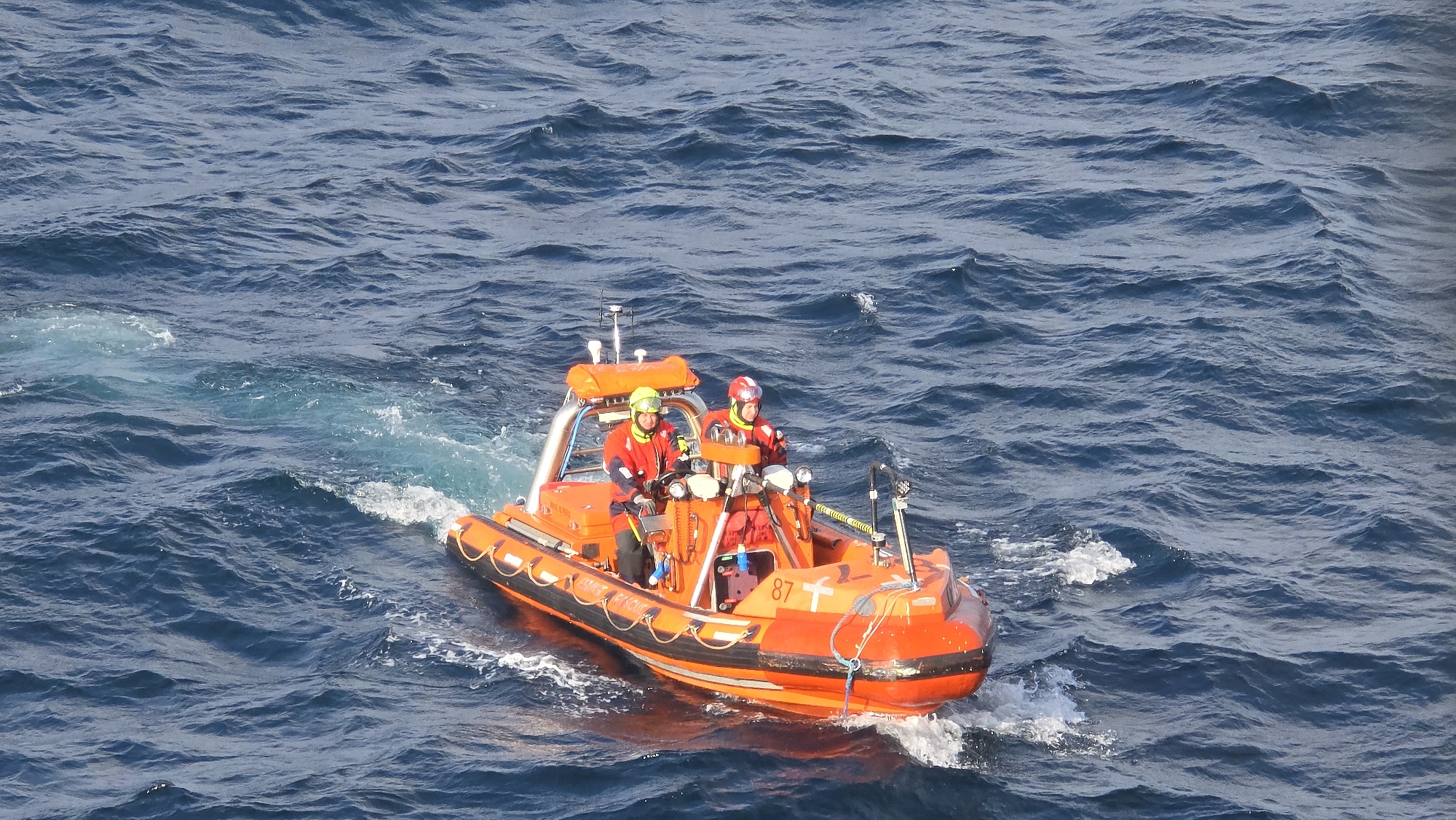 Two people wearing helmets and red suits are operating a bright orange inflatable boat with the number "87" on the front. The boat is moving through the water, creating a wake. The sea is dark blue with waves. The scene may be located off the coast of Randaberg, Norway, a coastal area, given the presence of the rescue boat.