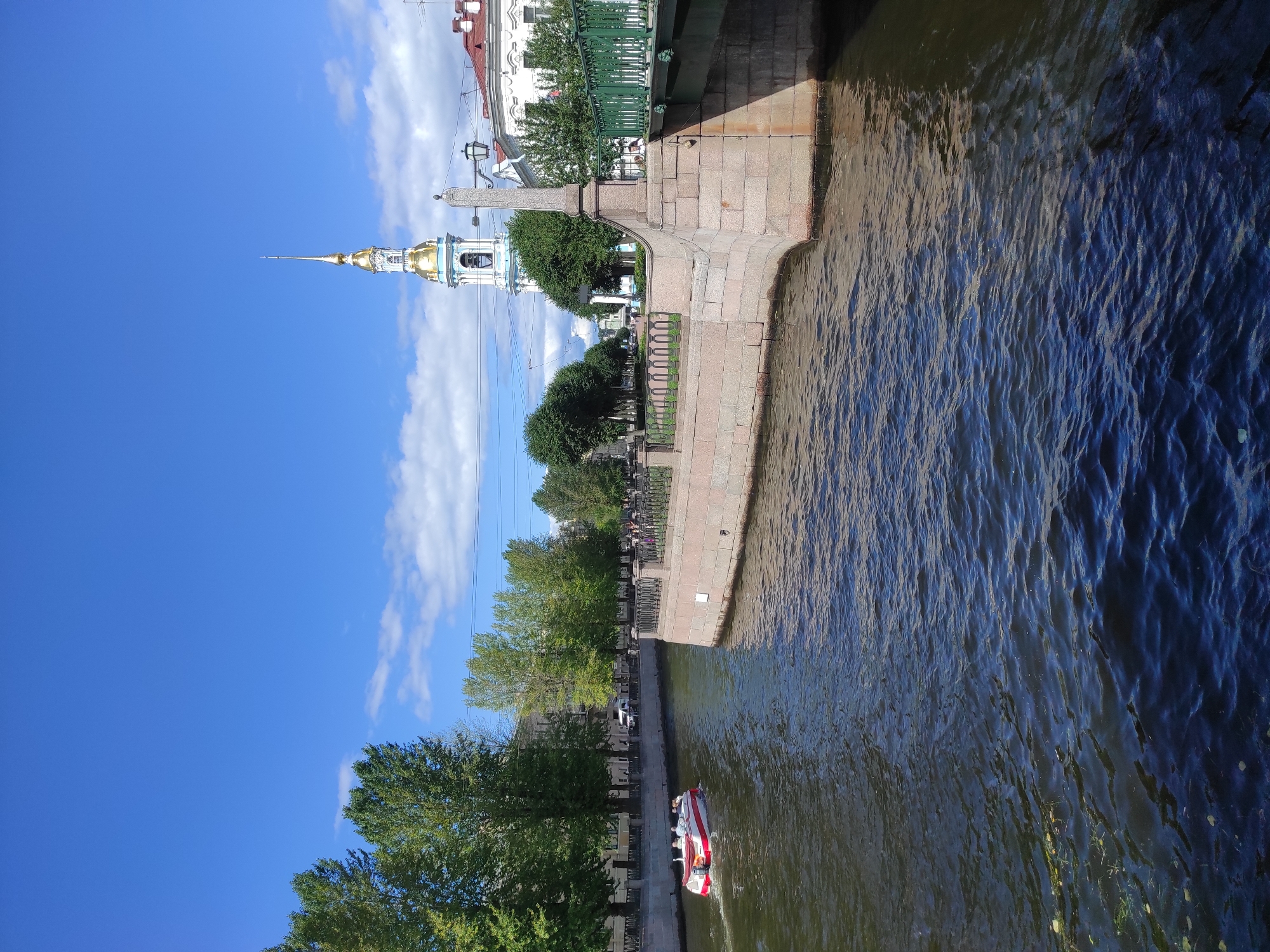 The image depicts a canal in Saint Petersburg, Russia, on a clear day. The canal's dark, rippling water fills the foreground. A white and red tour boat with several passengers is navigating the canal. On the right bank, a stone embankment is topped with a decorative railing and trees. Beyond the embankment, a bridge with a green railing is visible, leading to buildings. In the background, a tall, white and blue bell tower with a golden spire stands prominently against the blue sky. Lush green trees line the left bank of the canal, with buildings visible behind them. The scene conveys a peaceful urban atmosphere.