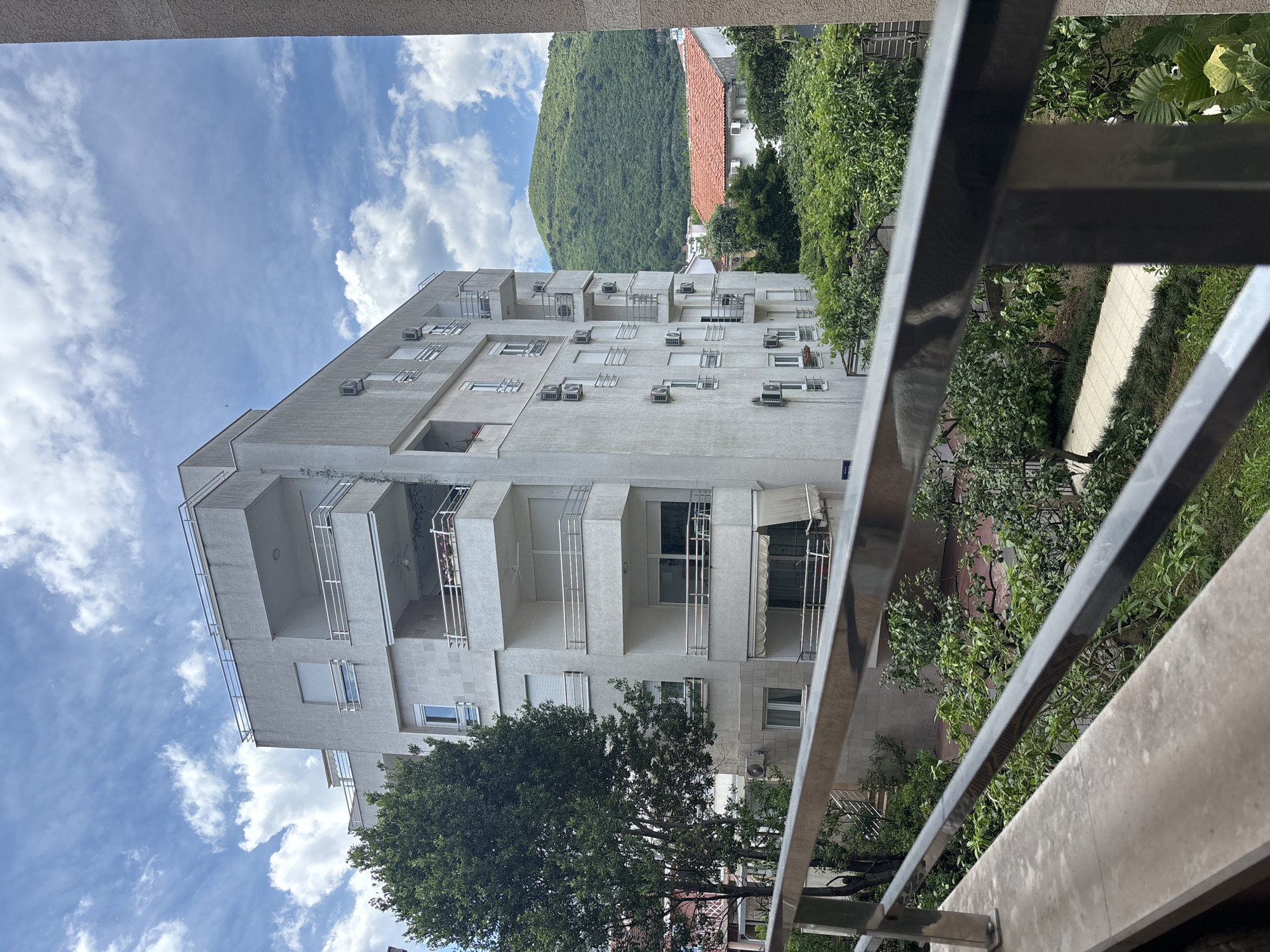 The image depicts a multi-story, light-colored building in Budva, Montenegro, with balconies and numerous air conditioning units mounted on its exterior. The building is viewed from an elevated position, likely another balcony, as a metal railing is visible in the foreground. Lush green trees and vegetation partially obscure the lower portion of the building and surround a sidewalk. In the background, a hill covered in dense vegetation is visible under a partly cloudy sky. A building with a red roof is partially obscured by trees. The overall impression is that of a residential area in a coastal town. There are no people visible in the image.