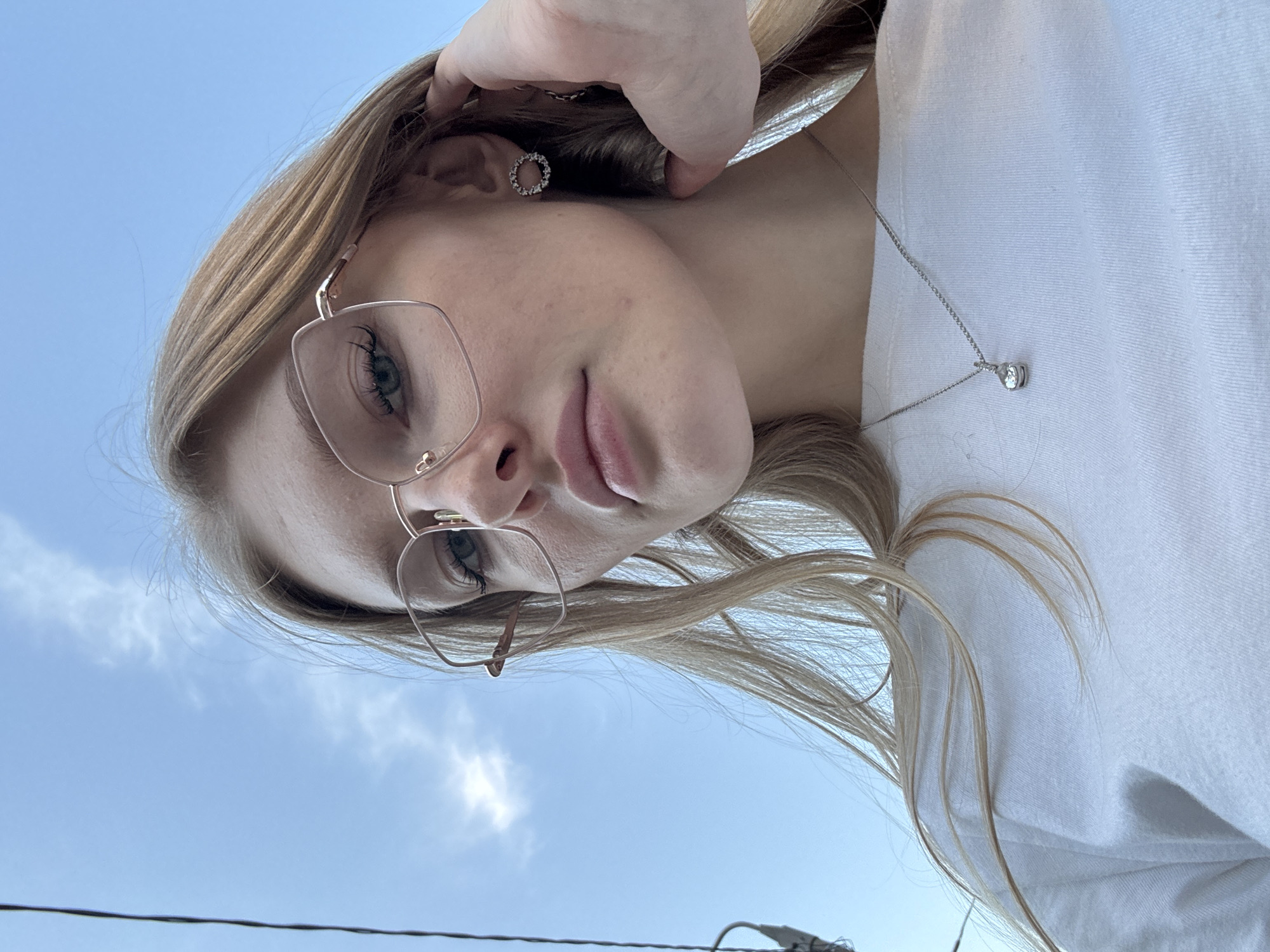 A young fair-skinned woman with blonde hair is looking down at the camera. She is wearing glasses and a white shirt, and a silver necklace with a small diamond pendant. She has a silver earring on her left ear. Her left hand touches her hair. The background is a blue sky with clouds. It is not possible to confirm the location as Perm, Russia from visual information.