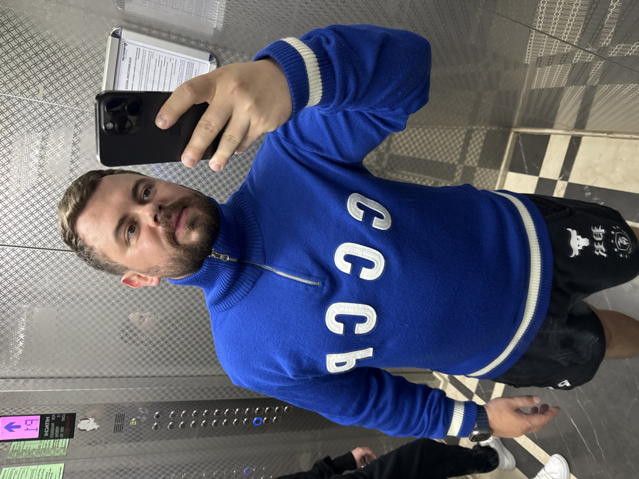 A man takes a selfie in an elevator. He wears a blue, zip-neck sweater with white trim and large white letters spelling "CCCP." Black shorts with a logo are visible, and he holds a dark-colored smartphone. The elevator interior features patterned walls and a tiled floor with a black and white checkerboard design. A control panel and signage are also visible in the elevator.