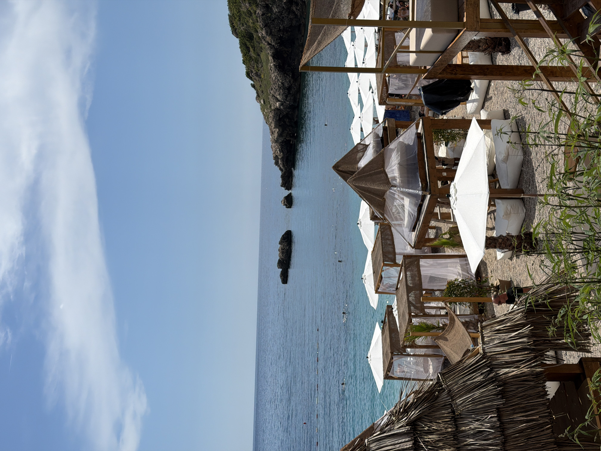 The image shows a beach resort in Drobnići, Montenegro. Several covered daybeds, umbrellas, and lounge chairs are set up on a pebble beach. The turquoise waters of the Adriatic Sea are visible in the background, with a rocky coastline and green vegetation to the right. A few people are swimming in the sea and sunbathing. The sky is blue with scattered clouds.