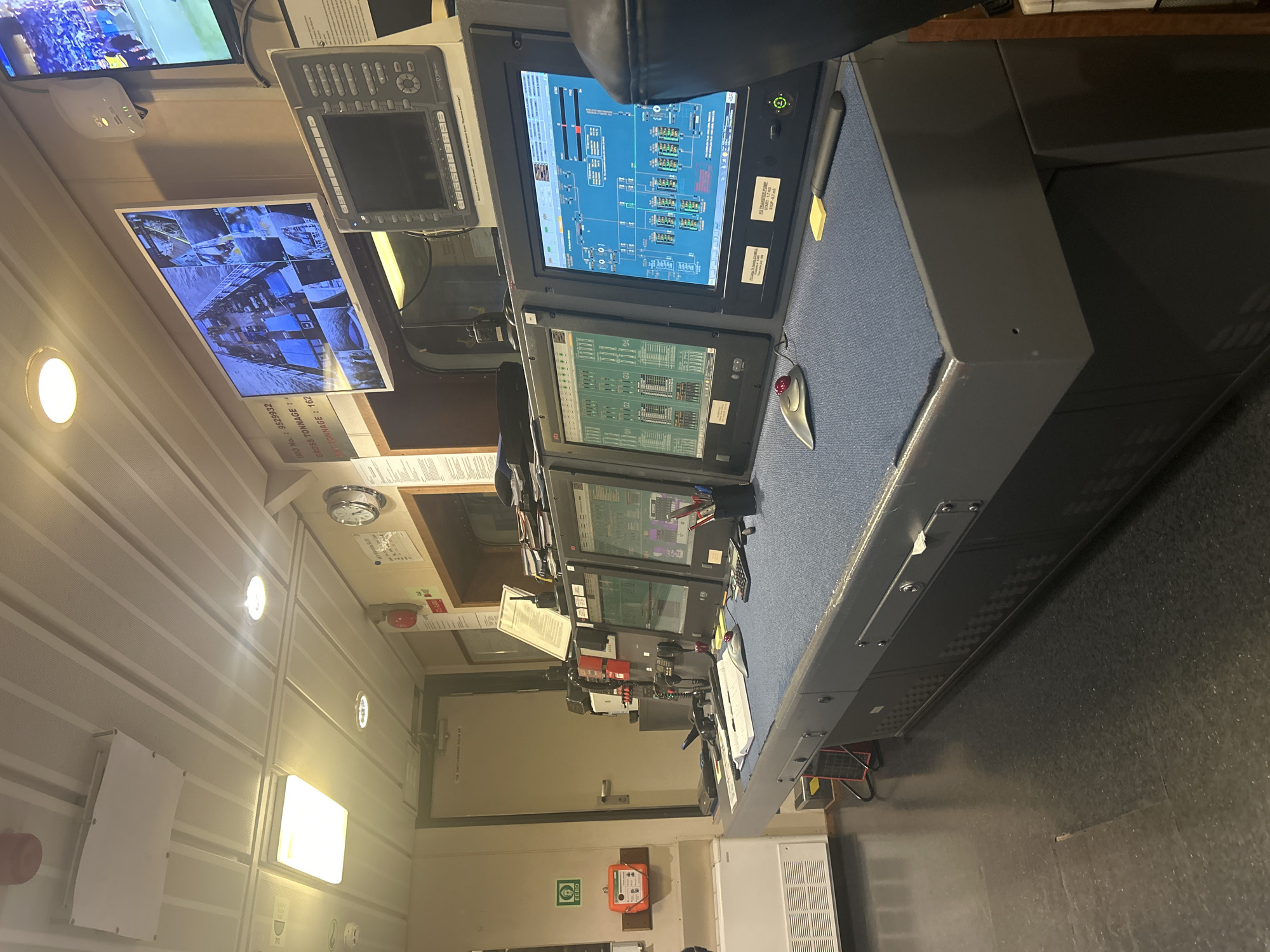 The image captures a room that appears to be a control center, possibly within a vessel or industrial facility. The focus is on a large console featuring multiple computer monitors displaying various diagrams and data visualizations. These monitors are arranged in a tiered setup, suggesting a hierarchical information display. Several input devices, such as a mouse and a stylus, are present on the console's surface. Above the console, a wall-mounted monitor shows a security camera feed displaying multiple views. The room itself is illuminated by recessed ceiling lights and appears to be located in a setting with functional and practical design. Text on the wall indicates details about the vessel and safety equipment location. An emergency breathing device and a door are present on the back wall. The overall impression is that of a functional and utilitarian workspace.