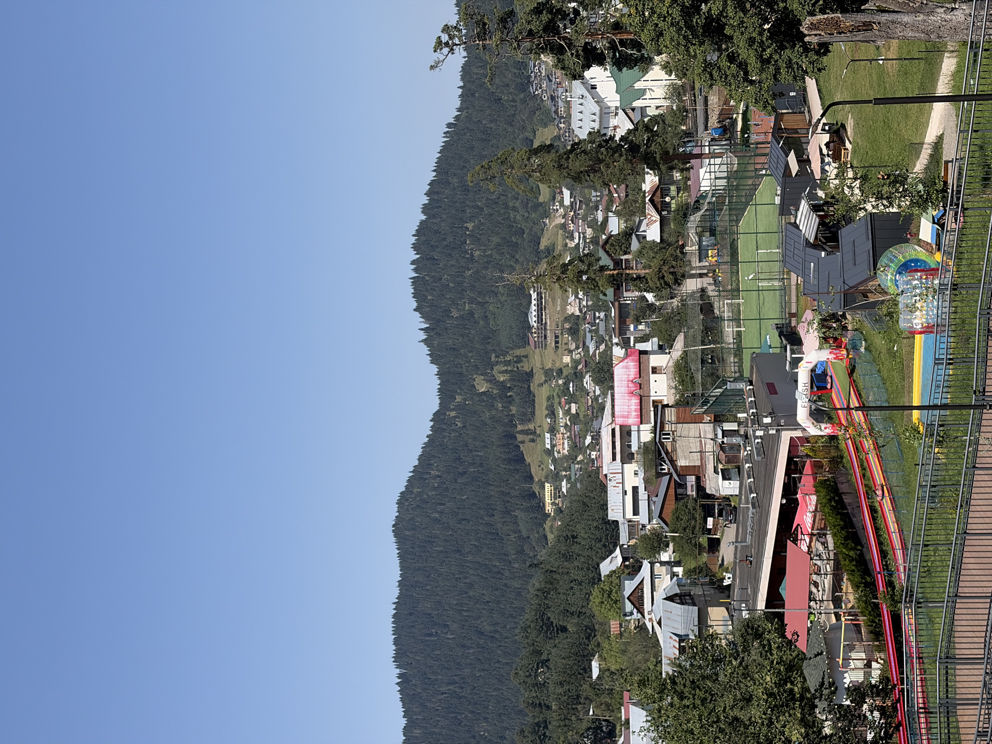 The image shows a daytime, elevated view of a village nestled in a valley at the base of a densely forested mountain range. The village consists of numerous buildings with varied architectural styles, including houses with metal roofs and some larger, modern structures. In the foreground, there is a recreational area with a sports field, possibly a soccer field, a colorful slide, and inflatable attractions. A prominent arched entranceway with the word "FLASH" is visible near the slide. The sky is clear and blue. The scene appears to be a resort or tourist area in Bakuriani, Georgia, given the mountainous terrain and the recreational facilities. There are no discernible people in the image.