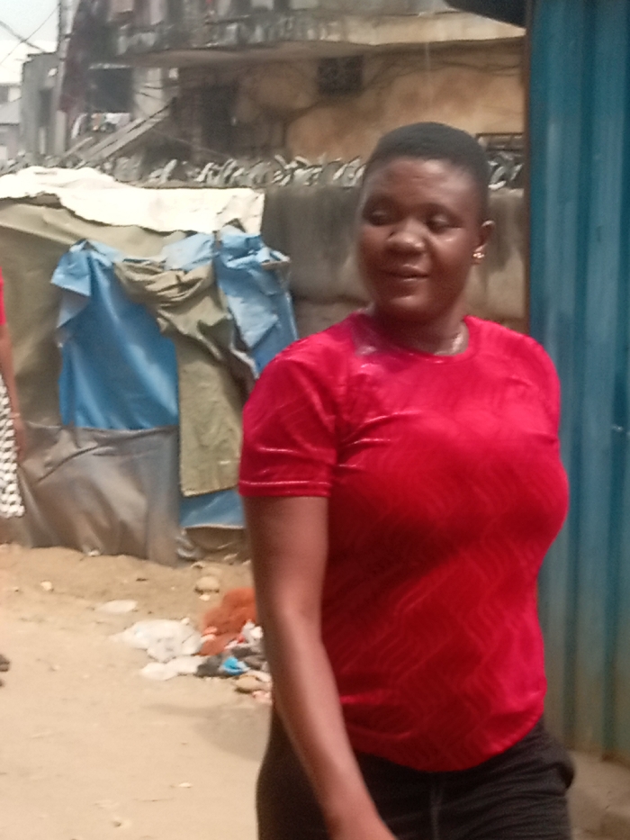 The image depicts an outdoor street scene in Atani, Nigeria. A woman with short, dark hair and light-brown skin occupies the central and right foreground. She wears a short-sleeved red top with a subtle textured wavy pattern and dark bottoms. A small gold-colored hoop earring is visible in her right ear. Her head is turned slightly to her right, and her gaze is directed away from the camera. A partial view of another individual, wearing a black and white patterned garment, is visible on the far left. The ground in the foreground consists of packed dirt with scattered debris, including plastic bags and fabric scraps. In the mid-ground, a structure composed of draped tarpaulins and fabric in beige, blue, and olive green is present. The background features multi-story concrete buildings, with a defensive barrier of broken glass fragments or sharp objects along a low wall. A vertical section of blue corrugated metal is visible on the far right.