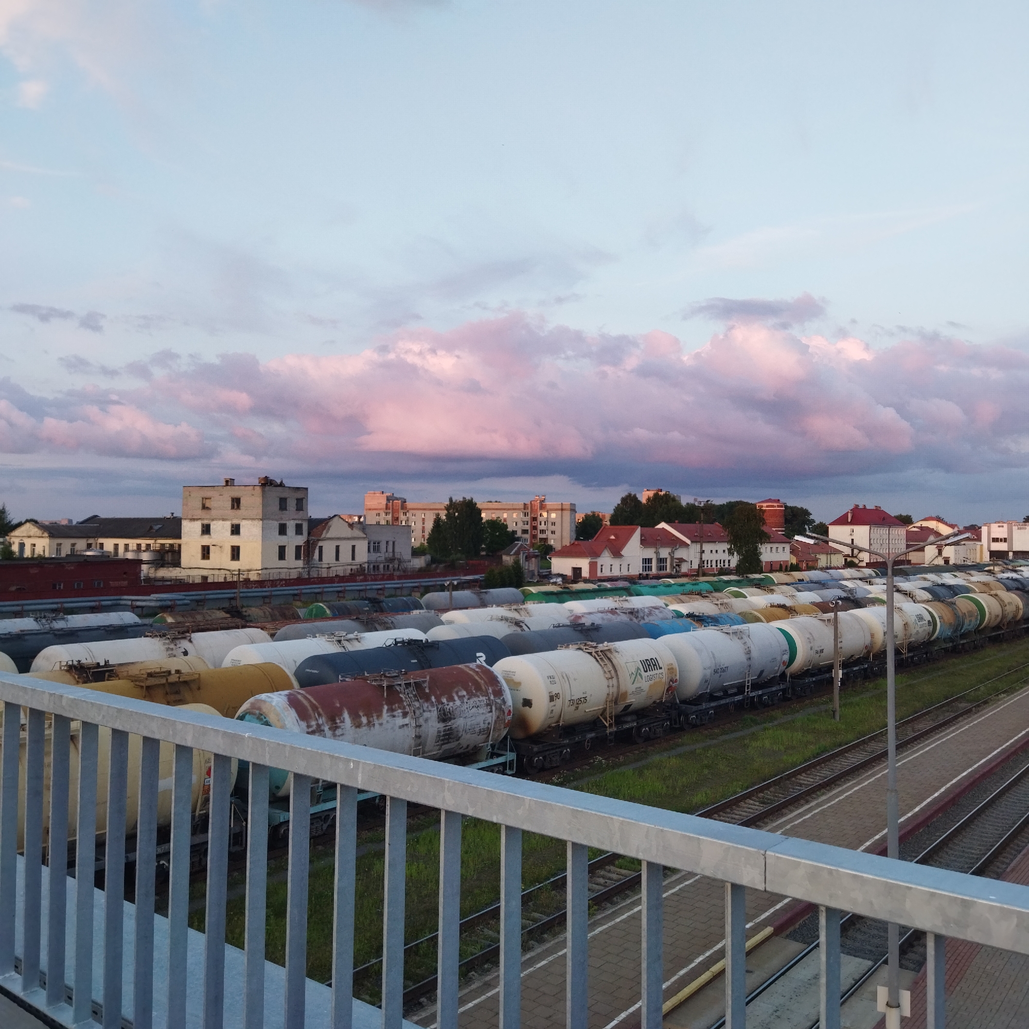 An elevated, eye-level, medium shot captures a vast train yard under a soft, pastel sky. The foreground is dominated by a silver metal railing, casting subtle shadows. Beyond the railing, numerous railroad cars, predominantly cylindrical tank cars in various colors like white, yellow, dark gray, and reddish-brown, are parked on parallel tracks. Some tank cars exhibit signs of rust. The middle ground reveals a collection of buildings, including industrial structures and residential buildings with red-tiled roofs, set against a backdrop of green trees. The sky is a canvas of pale blue with scattered clouds tinged with pink and purple hues, suggesting either dawn or dusk. The scene appears to be in a semi-urban or industrial area.