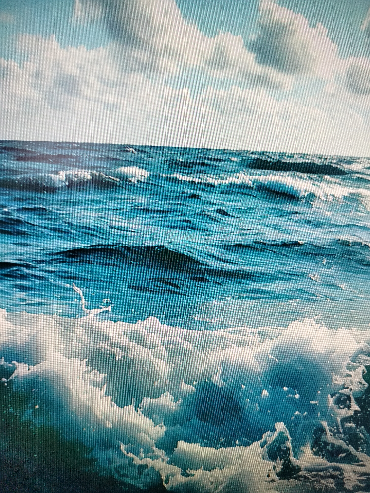 A close-up, eye-level shot captures a dynamic ocean scene under a partly cloudy sky. The water is a deep, rich blue, with white foam cresting on the waves, particularly in the foreground where a large wave is breaking. The sky above is a lighter blue, with scattered, fluffy white clouds. There are no people or man-made objects visible in the image, and no identifiable landmarks to suggest a specific location within Yashkino, Russia. The focus is entirely on the natural elements of the sea and sky.