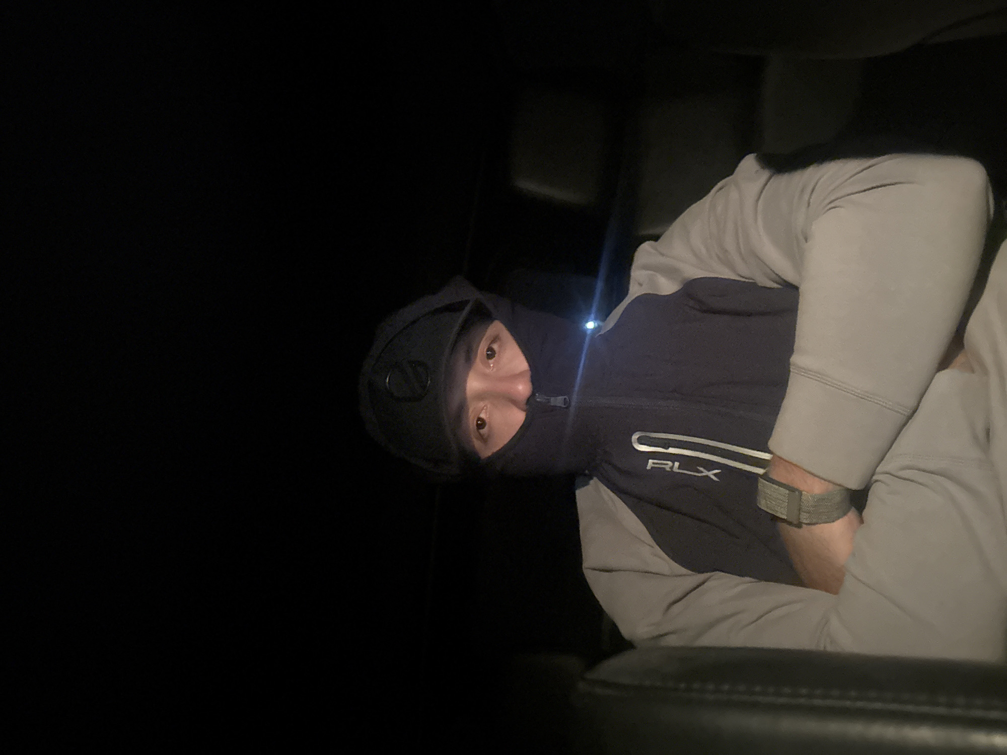 A person wearing a black balaclava and a black and grey RLX jacket is sitting in a dark car, with the front of the vehicle visible in the lower part of the frame. The location is unknown and cannot be inferred from the image.