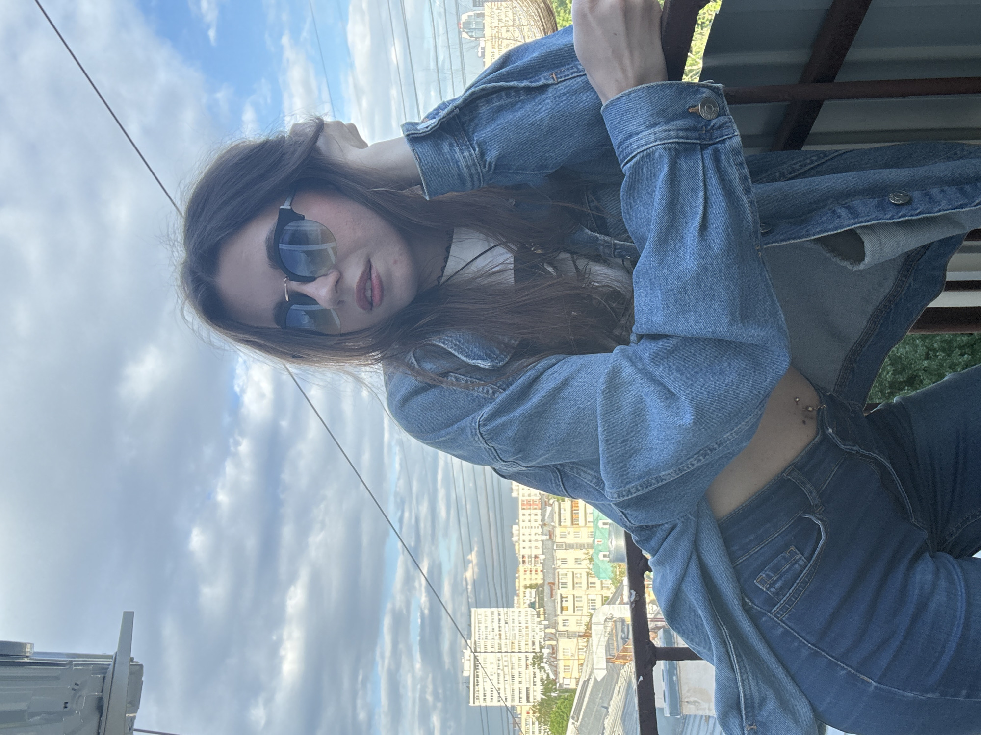 A young woman is posing outdoors. She is wearing a denim jacket over a white shirt, denim jeans, and sunglasses. She is standing on what appears to be a rooftop or high balcony, with a metal railing visible. In the background, there is a view of buildings, which suggests an urban setting. Given the architectural style of the buildings, it is plausible that this image was taken in Moscow, Russia. The sky is visible with scattered clouds. The photograph is rotated.