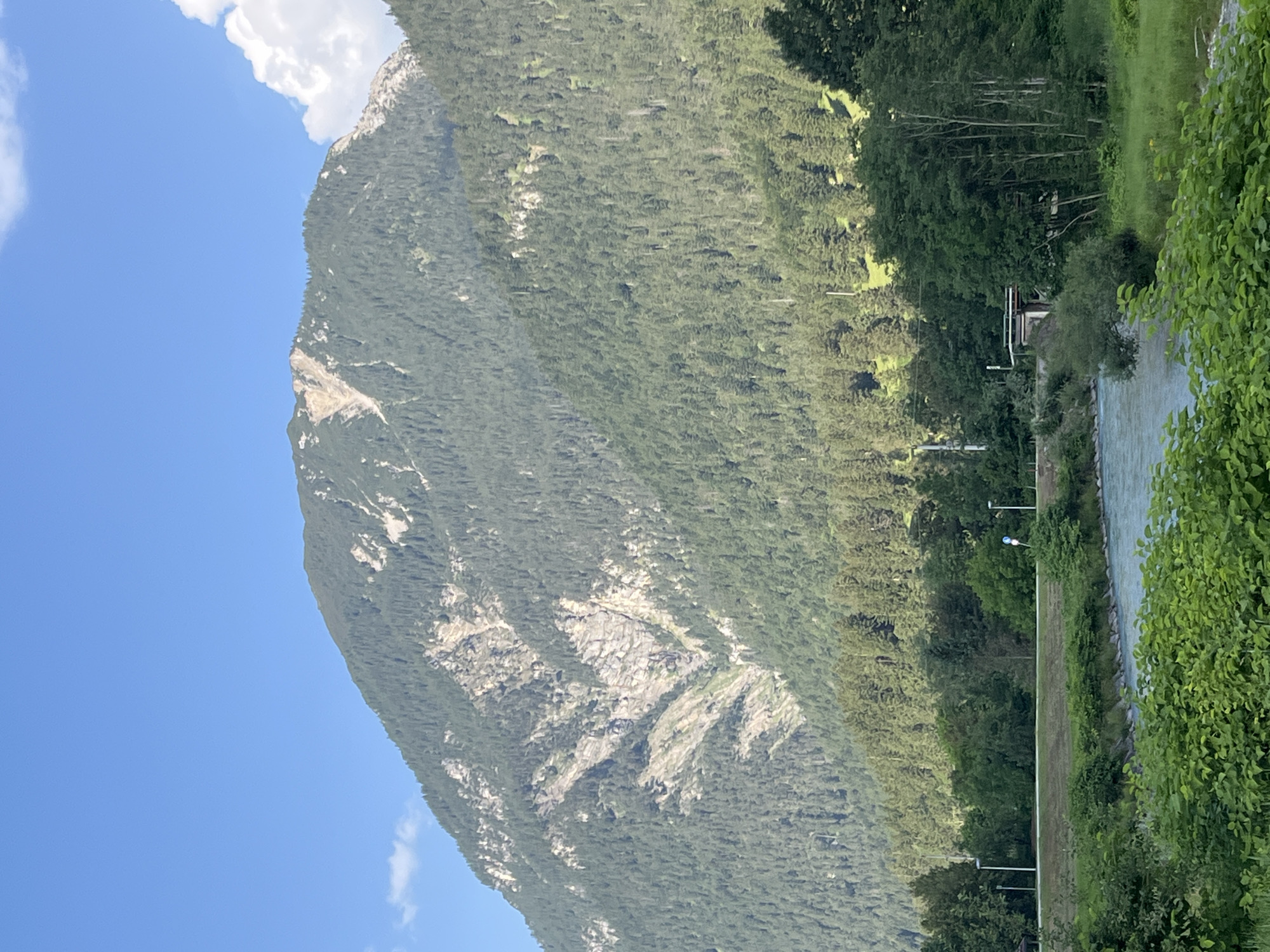 A serene mountain landscape is depicted. In the foreground, a clear blue river flows, bordered by lush green foliage. Beyond the river, a road runs parallel to the water, with a few sparse trees and some distant buildings visible. The middle ground is dominated by a densely forested mountain slope, transitioning into the majestic peaks of the Alps in the background. The mountain faces show a mix of dark green pine forests and lighter, rocky outcrops. The sky is a vibrant blue, with a few wispy white clouds. This scene captures the natural beauty of Garmisch-Partenkirchen, Germany, a region renowned for its stunning alpine scenery.