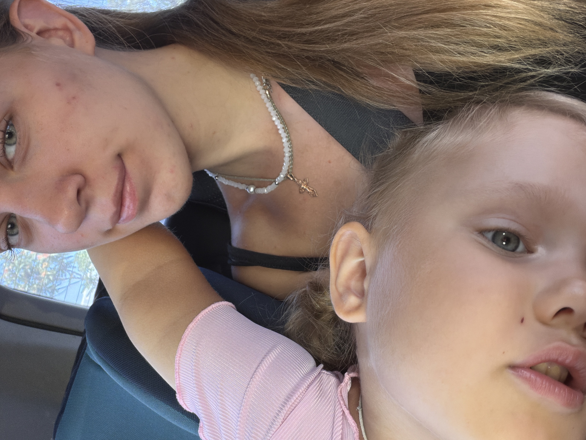 The media file shows a close-up, selfie-style shot of two individuals, likely children, inside a vehicle. The person in the foreground on the right appears to be a young child with blonde hair, their face tilted towards the camera, with their left eye and mouth visible. They are wearing a pink ribbed top. To the left and slightly behind, a second individual, possibly an older child or teenager, is also captured, with their head tilted and their left eye, cheek, and neck visible. They have long brown hair and are wearing a black top underneath a necklace with white beads and a small cross pendant. The background suggests the interior of a car, with a car seat visible in dark blue and a glimpse of trees and sunlight through a window, indicating it is daytime and likely sunny. The composition is informal, suggesting a candid moment or a spontaneous selfie. There is no visible text in the image. The overall mood appears casual and personal.
