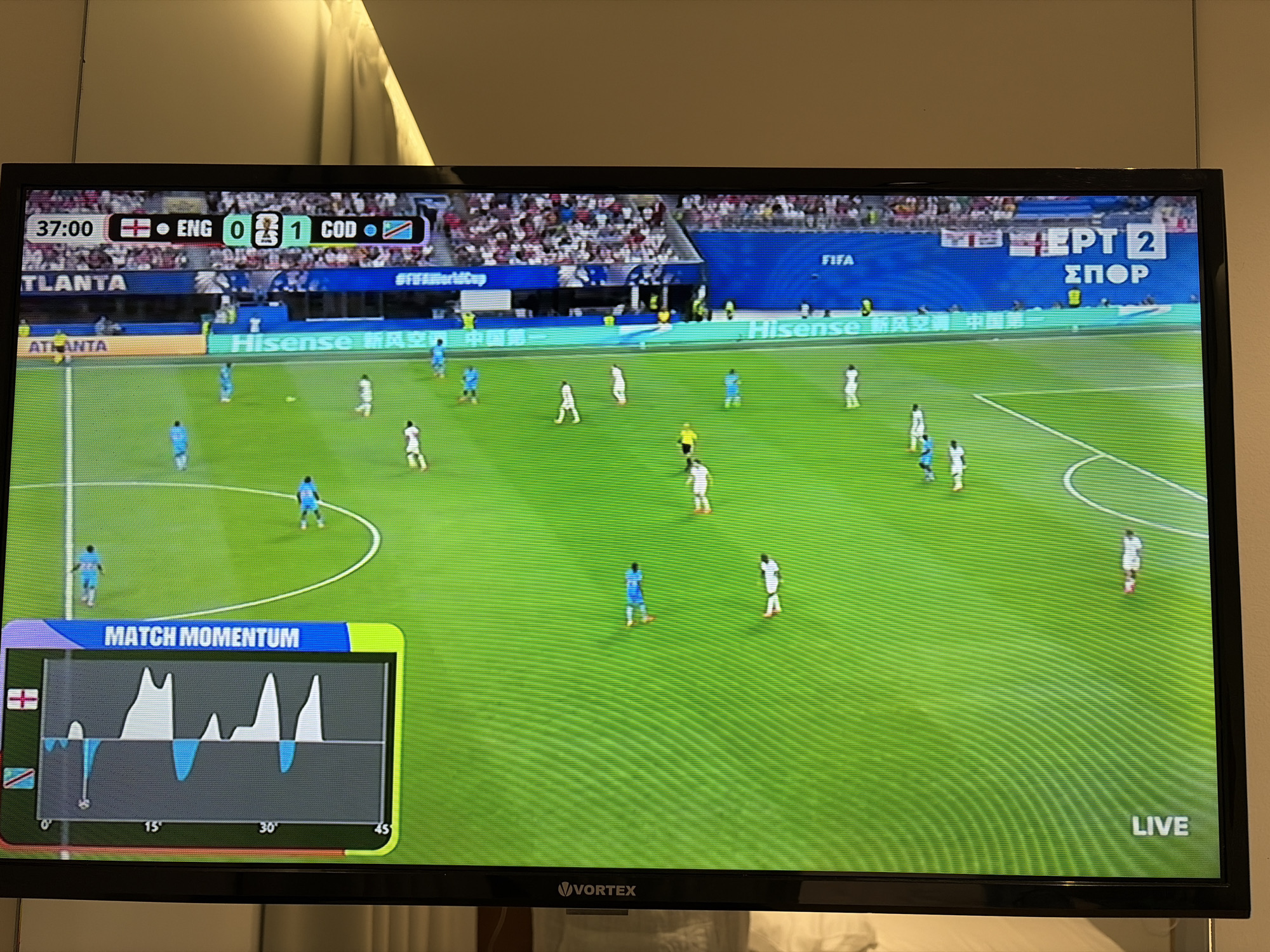 This is a medium shot of a television screen displaying a soccer match. The game is between England (ENG) and Congo (COD), with the score currently 0-1 in favor of Congo. The game clock shows 37:00 minutes elapsed. The scene is a brightly lit soccer stadium with green turf. Several players in white and blue uniforms are visible on the field. In the foreground, a graphic labeled "MATCH MOMENTUM" displays a line graph that fluctuates over time, with icons of the English and Congolese flags. The lower right corner of the screen shows the word "LIVE" in white text. The top right of the screen displays "EPT 2 ΣΠΟΡ", likely indicating the television channel. The bottom of the television frame shows the "VORTEX" logo. The overall setting appears to be indoors, with the television screen reflecting a wall with what looks like a diffused light source above it. The atmosphere is that of a live sporting event.