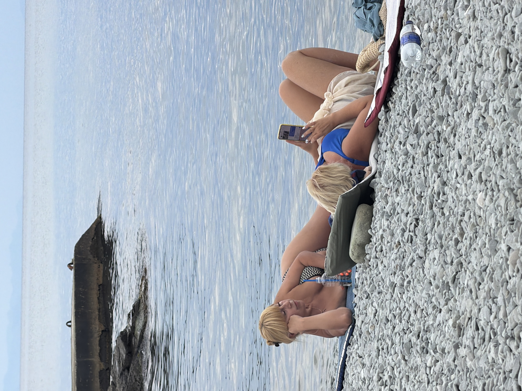 Two women are lying on a pebble beach next to calm water. The woman on the left, lying on her side with her head propped on her hand, is wearing a black and white patterned bikini. A blue bottle with a straw is near her. The woman on the right is lying on her back, looking at her cell phone. She is wearing a blue bikini top and a light-colored sarong. A straw hat and a folded blue cloth are next to her. In the background, a concrete pier extends into the water. The sky is overcast, and the water is a pale blue. The scene appears to be a peaceful day at the beach.