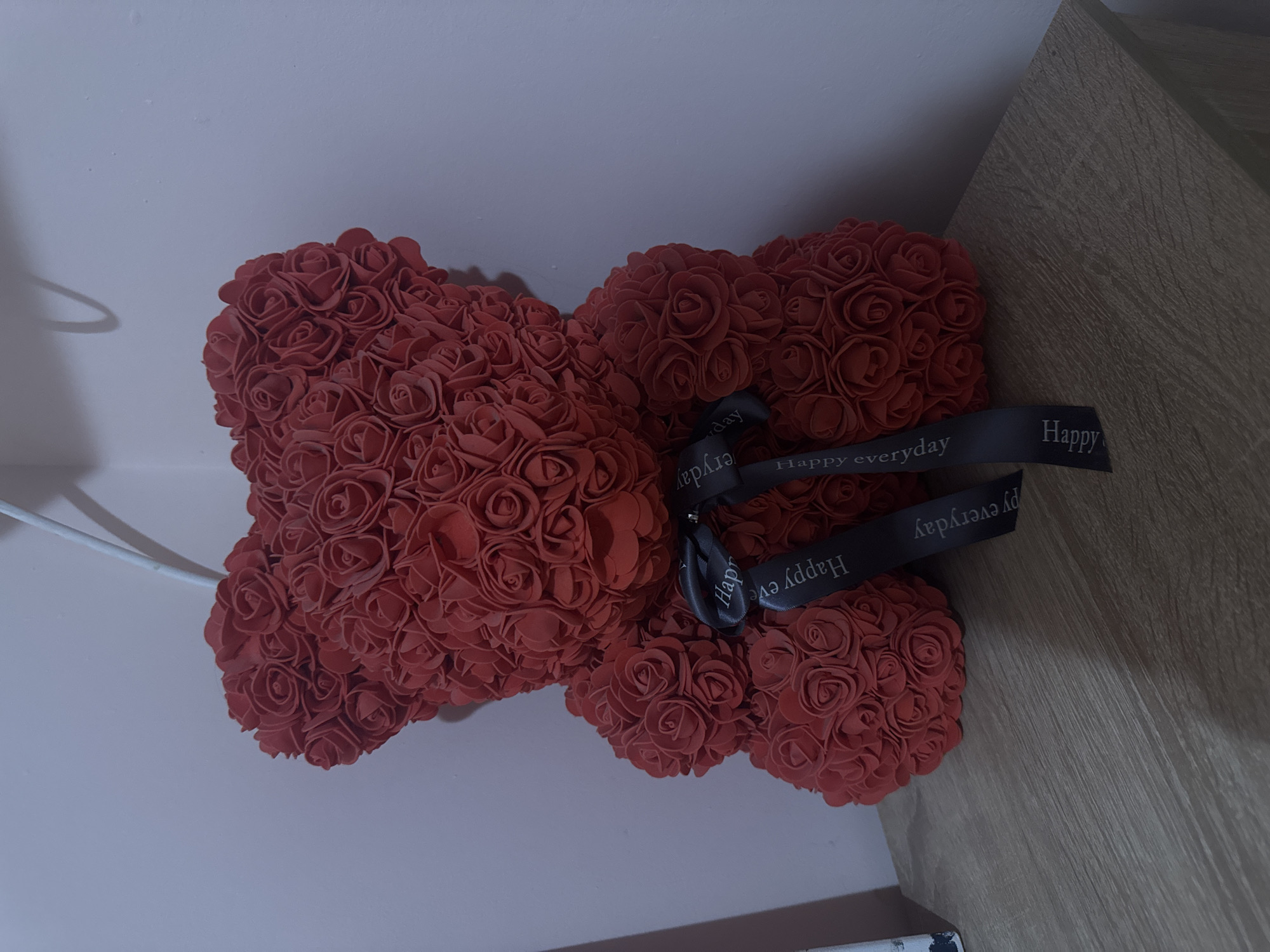 A red rose teddy bear with a black ribbon that says "Happy everyday" is placed on a table with a gray wall in the background. The location is not discernible from the image. No people are visible. The only object is the teddy bear. There are no actions or interactions taking place. The environment is a room with a gray wall and a table.