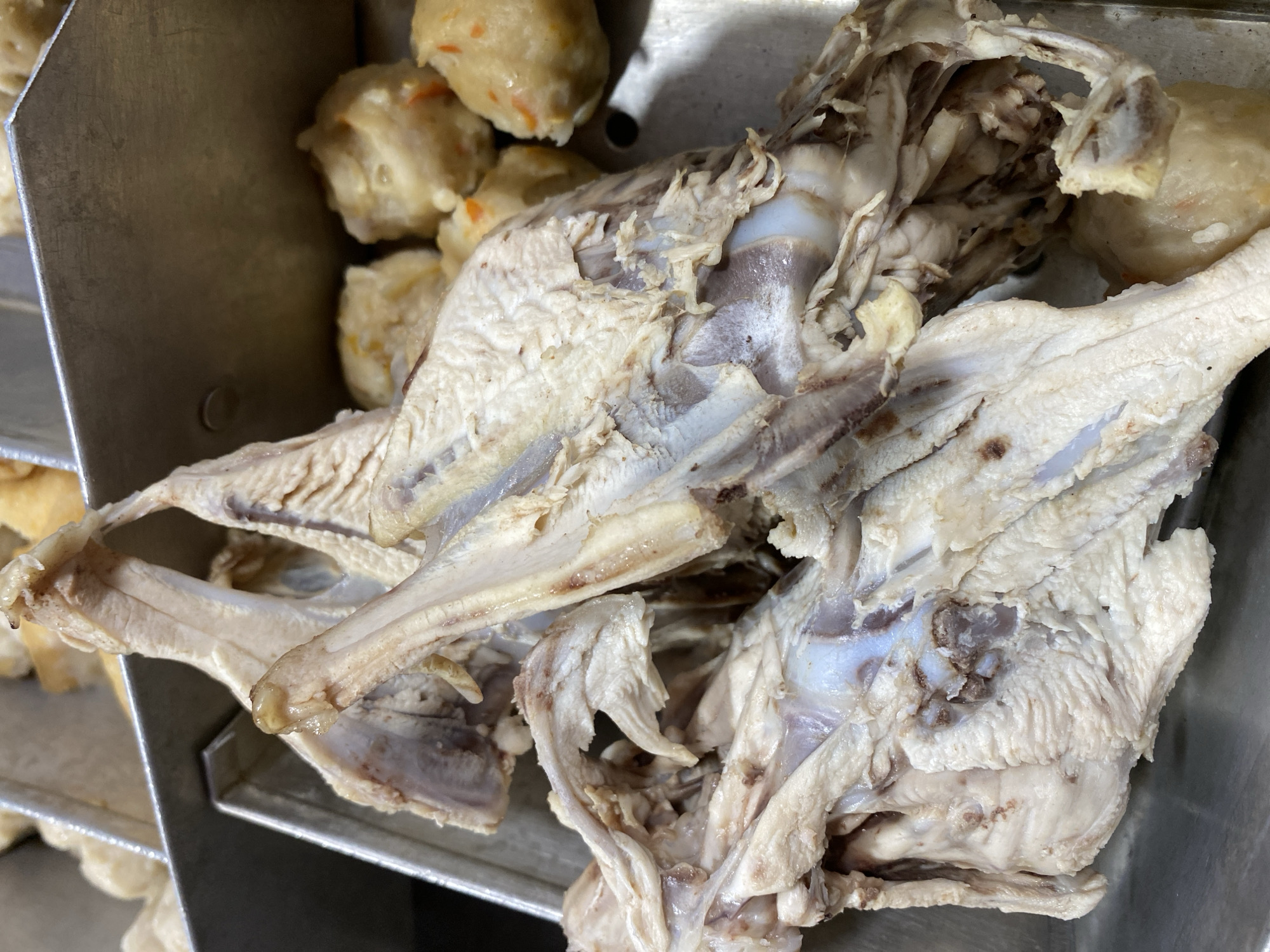 The image shows a close-up of a whole, cooked chicken carcass in a metal tray. The chicken is pale and appears to be boiled or steamed, with some skin and meat visible, but the bones and internal structure are exposed in many areas. The chicken is positioned towards the foreground and center of the image. In the background, to the right of the chicken, are several small, round, light-colored food items, possibly meatballs or dumplings, also in the metal tray. The environment appears to be a food preparation or serving area, likely in a market or restaurant. There are no people visible in the image. The image does not provide any information about the city of Kasihan, Indonesia.