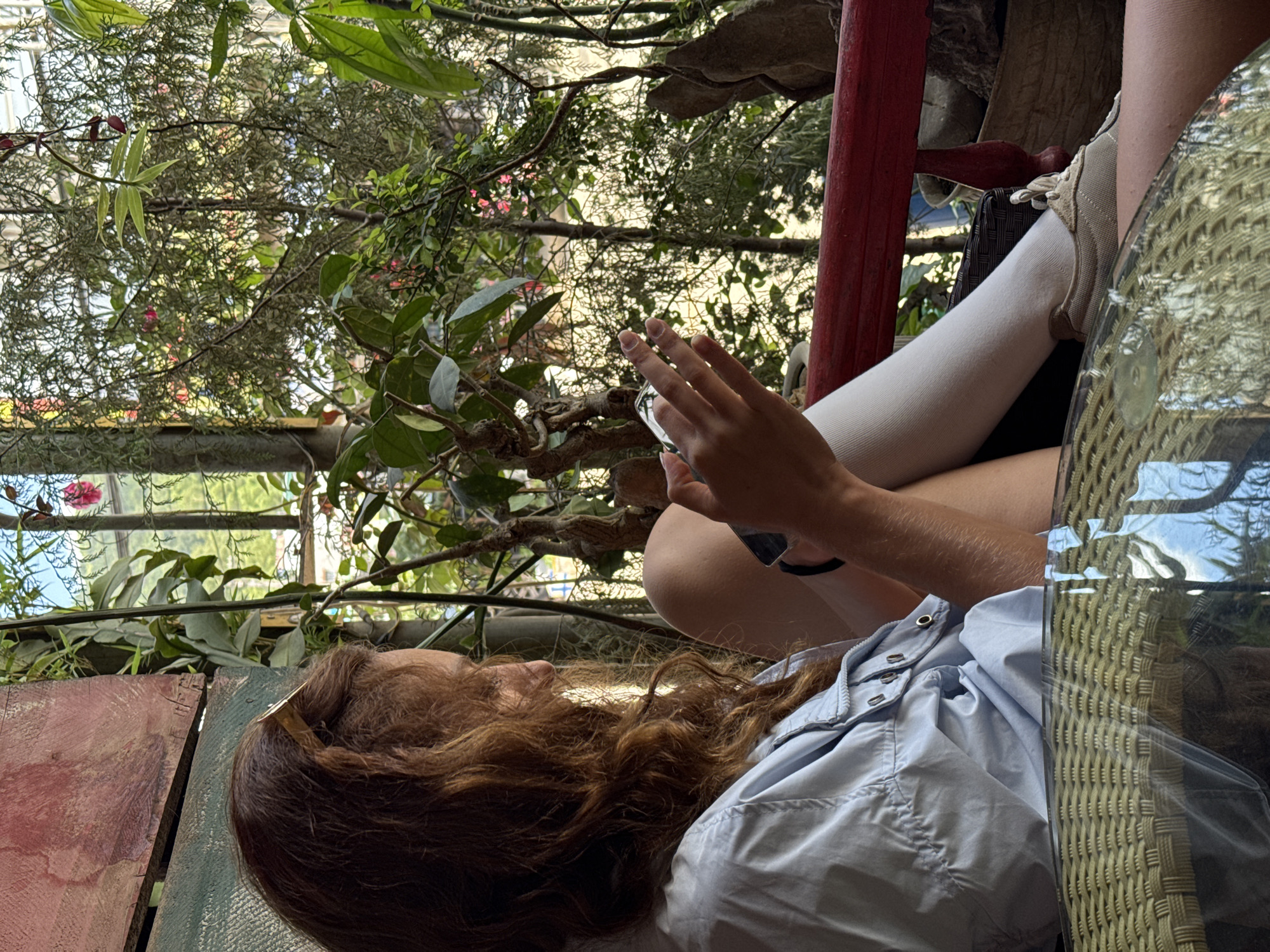 A person with long brown hair sits at a wicker table in what appears to be an outdoor seating area, potentially a restaurant or cafe. The location appears to be an area with abundant vegetation. The person wears a light blue shirt and white socks and appears to be using a mobile phone. The background includes trees, vegetation, and structures. The location is Đồng Văn Commune, Vietnam.
