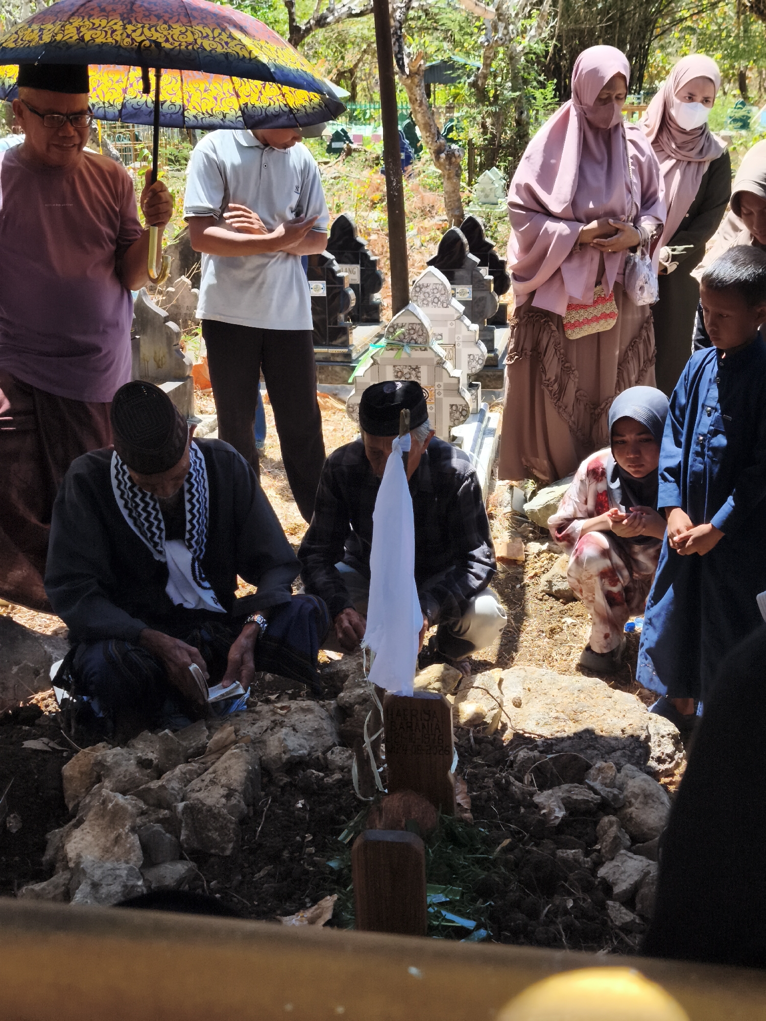The image depicts a funeral ceremony taking place outdoors in a cemetery in Tonasa, Indonesia. Several individuals are gathered around a grave. In the foreground, an elderly man wearing a black cap and scarf sits on the ground, holding what appears to be money or papers. Beside him, a young person in a headscarf and patterned clothing kneels, with hands clasped. A man in a dark shirt and cap is also present, looking down. Behind them, a man holding a colorful umbrella stands over a person in a light-colored shirt. Further back, two women in headscarves and modest attire are observing the proceedings. The gravestones are visible in the background, along with trees and foliage, suggesting a sunny day. The overall atmosphere appears somber and respectful.