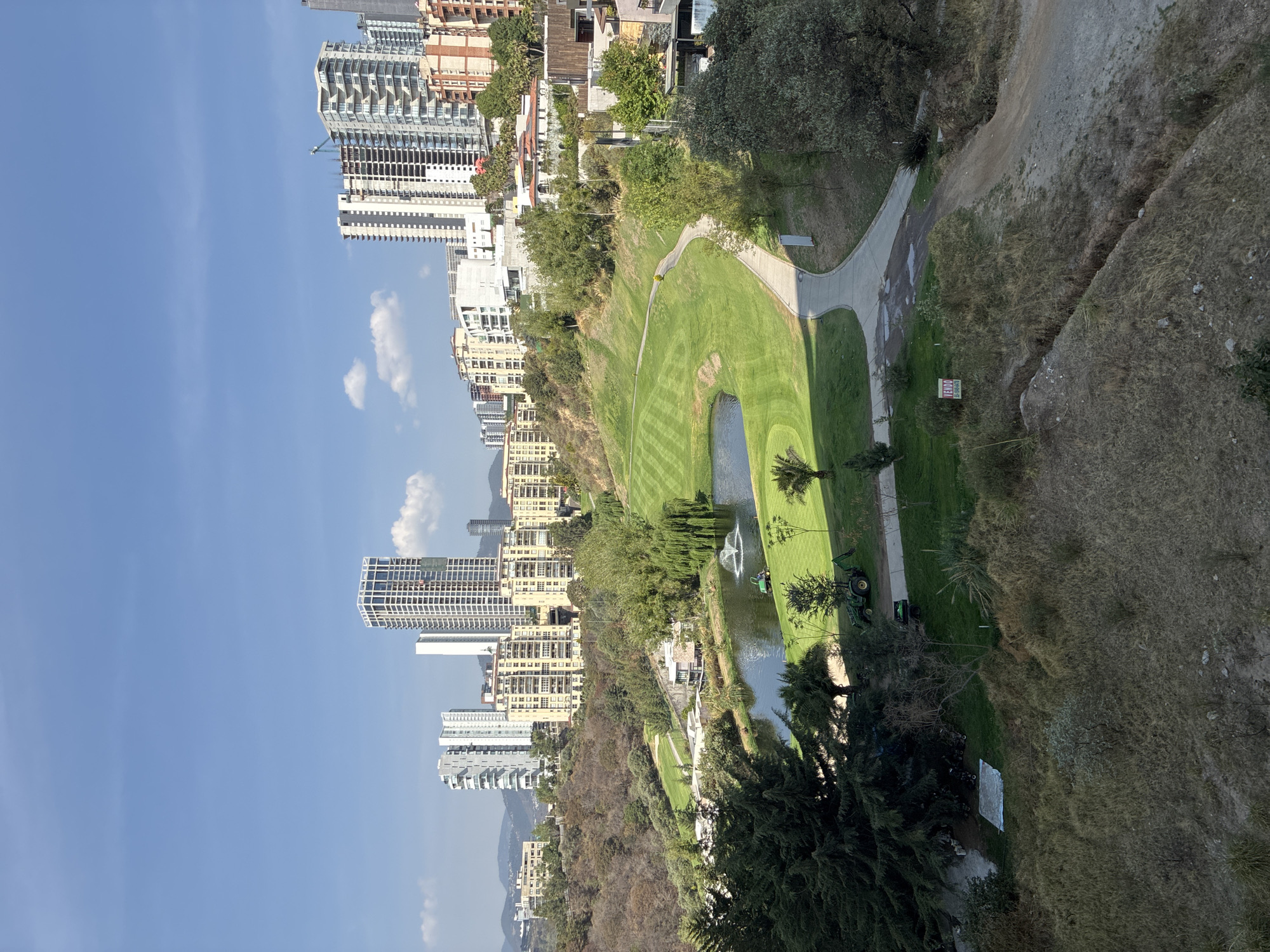 The image captures a panoramic view of an urban landscape seamlessly integrated with a vibrant green recreational area, under a clear blue sky. On the left and stretching across the background, numerous modern high-rise buildings dominate the skyline, some appearing to be residential with multiple balconies. Several structures are visibly under construction, indicated by exposed frameworks and the presence of a construction crane. Distant mountains or hills are visible on the far horizon.
In the midground, an expansive, meticulously maintained green space resembling a golf course occupies the central area. The grass features distinct striped patterns, and a tranquil body of water, possibly a pond, includes a fountain actively spraying water. Winding paved paths cut through the green lawns. A green John Deere tractor and a green golf cart are parked on the grass near the pond, suggesting maintenance activities. Various trees, some tall and mature, line the edges of the green space, separating it from the urban development and the wilder terrain.
To the right, the landscape transitions into a more natural, rugged environment with dry, light brown grass, sparse vegetation, and dirt slopes. A narrower dirt path is visible ascending a hill. A small red and white sign stands on the right, displaying Spanish text that reads "EMERGENCIAS" (Emergencies) and "OCUPADO" (Occupied). The time of day is daytime, evidenced by the bright, clear blue sky with a few scattered white clouds. No people are visible in the scene. The setting blends urban development with natural and recreational zones, typical of an area like Huixquilucan de Degollado, Mexico.