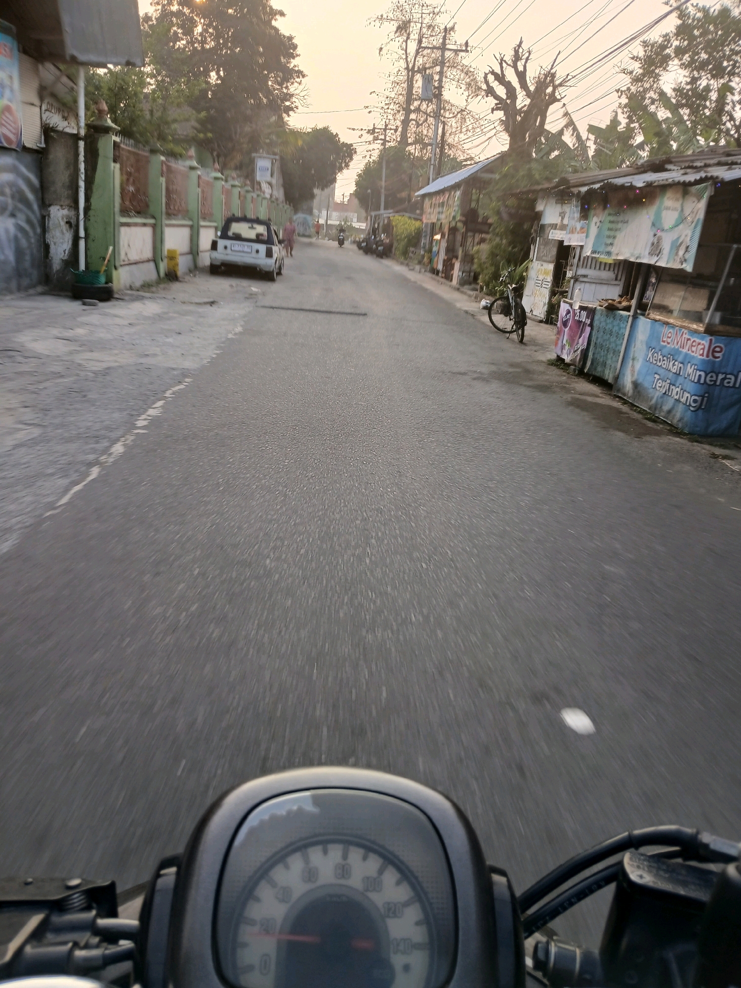 This is a first-person perspective, forward-facing shot taken from a motorcycle or scooter on a road in Depok, Indonesia. The scene depicts a street with buildings and trees lining both sides. The time of day appears to be late afternoon or early evening, as indicated by the warm, soft lighting and long shadows.
On the left side of the road, there is a wall with a green trim and a rusted metal fence. A white car is parked on the side of the road. Further down the street, a person in pink clothing is walking away from the camera.
On the right side of the road, there are several small shops or stalls with corrugated metal roofs. One stall has a large blue banner with text that appears to be advertising "Le Minerale" bottled water. A bicycle is parked in front of the stalls.
The road surface is asphalt, and there are power lines and utility poles overhead. The foreground shows the instrument panel of the motorcycle or scooter, with a speedometer visible. The overall impression is a quiet, everyday street scene in an Indonesian city.