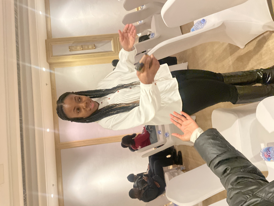 A woman with braided hair, wearing a white blouse, black pants, and black boots, stands in a room with beige walls and white chairs. She smiles, holding something in her raised hand. Other people sit in the chairs behind her. The seating arrangement is such that people are sitting close to each other. A hand reaches out in front of the woman. Water bottles are sitting behind many of the chairs.