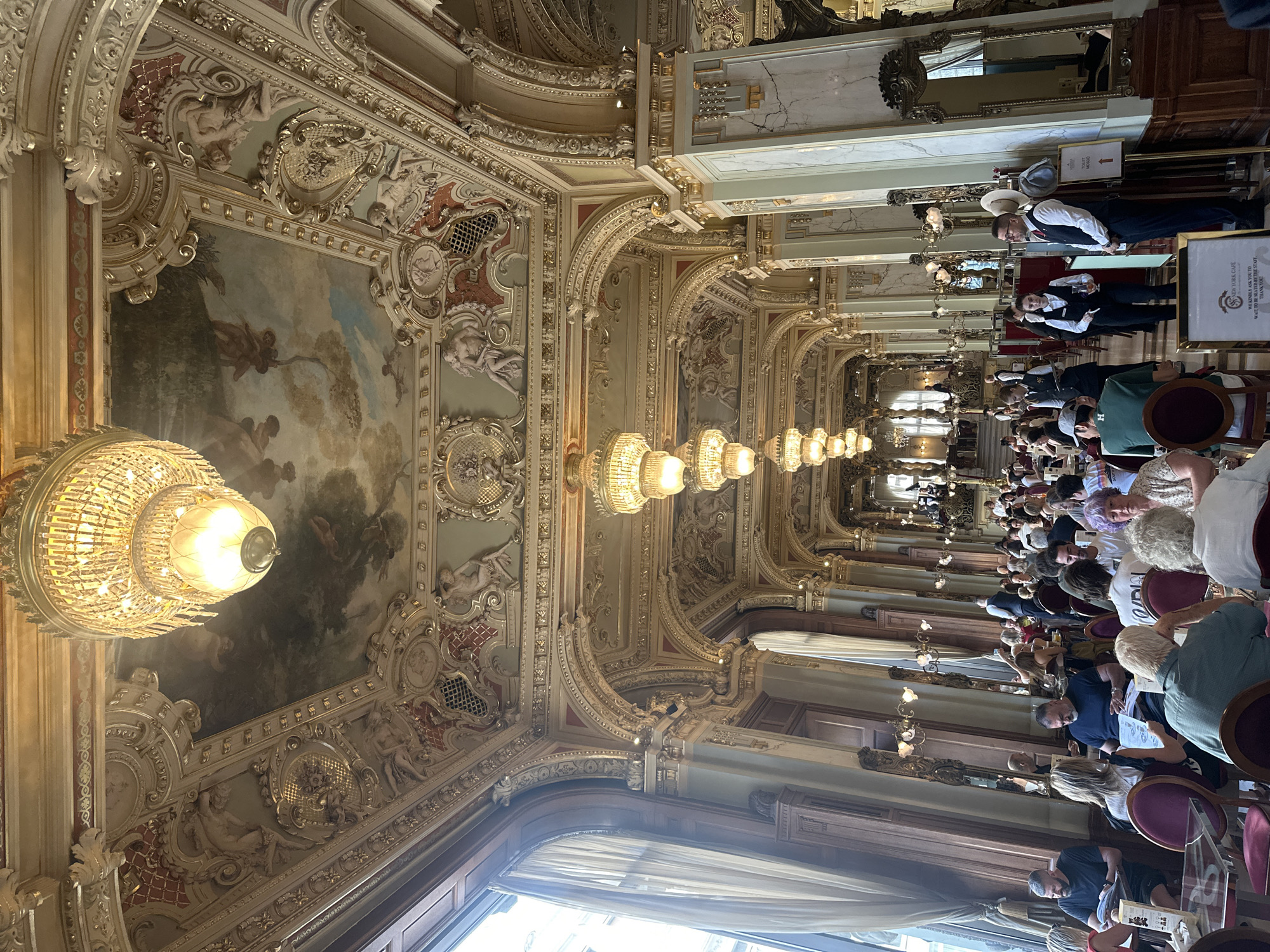 This image shows the interior of the New York Café in Budapest, Hungary. The café is elaborately decorated with ornate gold trim, frescoes on the ceiling, and crystal chandeliers. Patrons are seated at tables, many of whom are engaged in conversation or reading. Waitstaff in uniforms are visible, attending to the customers. The architecture suggests a historic and luxurious establishment, likely a popular tourist destination. Natural light streams in from large windows, illuminating the opulent interior.