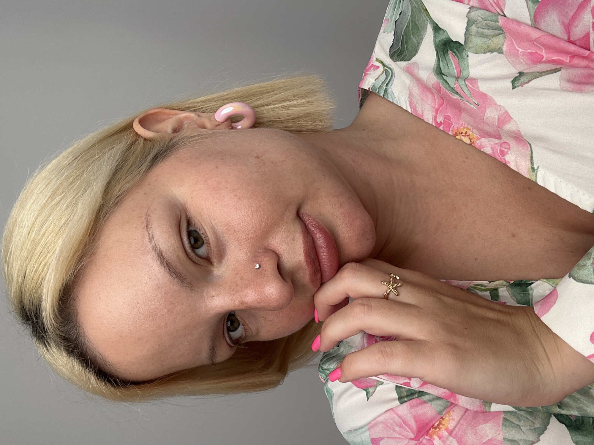 A close-up portrait shows a woman with blonde, chin-length hair, her head tilted and gaze directed towards the viewer. She has light brown eyes and a small silver stud piercing on her left nostril. Her lips are full and slightly parted, with a soft, natural color. Her chin rests gently on her right hand, which has bright pink nail polish and a gold-toned starfish-shaped ring on the ring finger.
She is wearing a white garment with a vibrant floral pattern featuring large pink blooms and green leaves. A light pink, C-shaped earring or ear cuff is visible on her left ear. Her skin appears fair with some freckles visible across her nose and cheeks. The background is a plain, solid light gray, suggesting an indoor setting with soft, even lighting. The woman's expression is calm and somewhat posed. No text is visible in the image. The location context for this image is Budva, Montenegro.