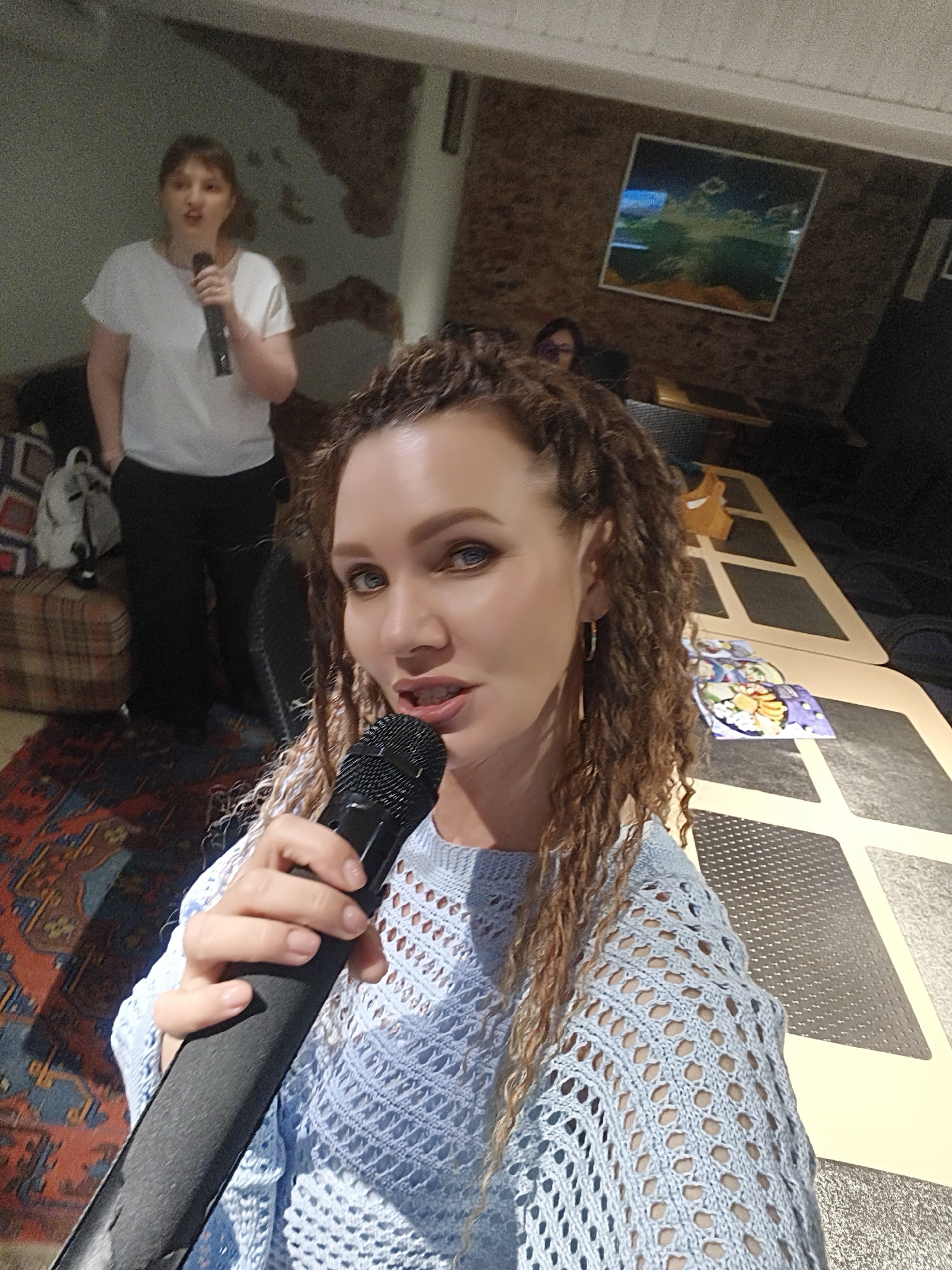 A woman with curly, light brown hair, wearing a light blue, lacy sweater, is holding a black microphone and looking directly at the camera. Behind her, another woman stands, also holding a microphone, and wearing a white t-shirt and black pants. A plaid couch is to the left of the second woman, and a white backpack sits on the couch. In the background, there is a long table with geometric designs and what appears to be a picture hanging on the wall. The location is Unknown, Unknown.