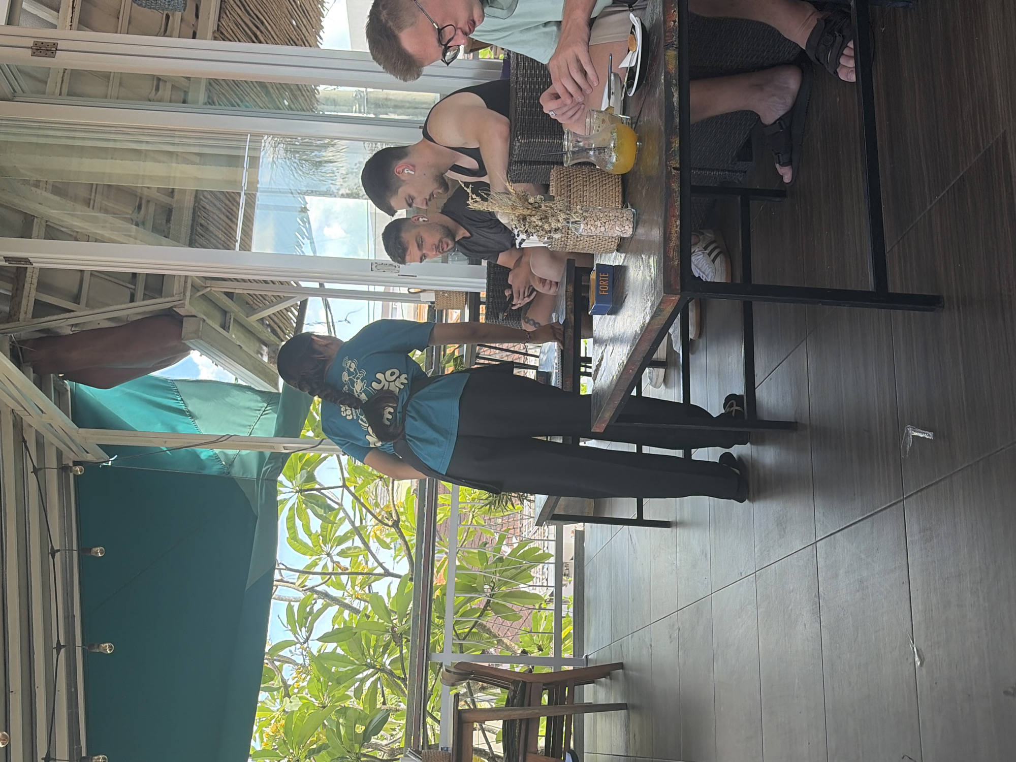The image depicts a scene at an outdoor cafe in North Kuta, Indonesia. A female server, wearing a teal t-shirt and black pants, stands facing away from the camera, holding a small dark object. In the foreground, a wooden table has a pitcher of orange liquid, a small basket with dried decorative reeds, a blue box with the word "FORTE" on it, and a black saucer with a small pastry or cookie. To the right of the table, a man wearing glasses is visible, partially obscured. Behind him, two men sit at a table, both wearing wireless earbuds. One man in a black t-shirt holds a phone. The other man wears a black tank top. The cafe appears to be an open-air establishment with a teal awning and railings visible. Lush green foliage and palm trees are seen in the background, suggesting a tropical environment. The floor is tiled.