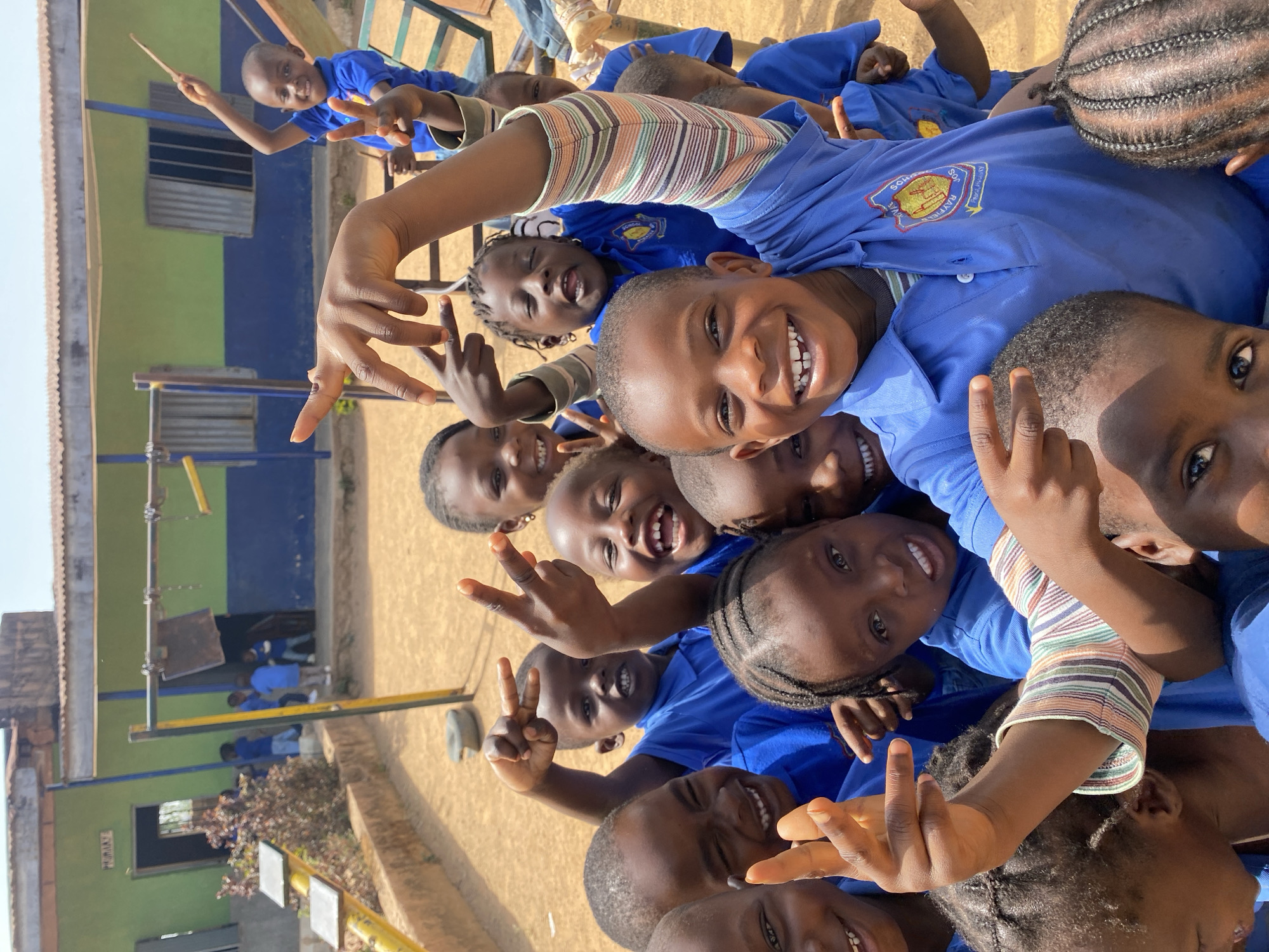 A group of children in blue school uniforms are tightly grouped together, smiling and making peace sign hand gestures towards the camera. The background shows a building with green and blue walls, a playground structure, and brown, sandy-looking ground. This photograph was taken in Gura Top, Nigeria.