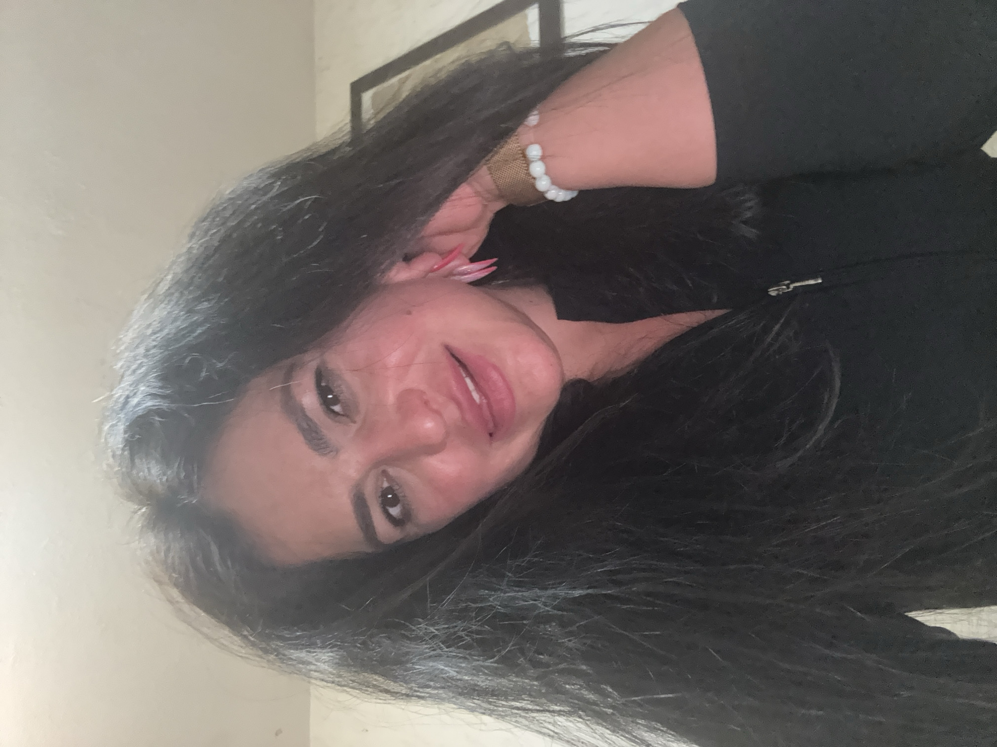 A woman with long, dark hair is shown in a close-up, angled portrait. She is wearing a black garment, possibly a zip-up jacket. A light-colored beaded bracelet adorns her wrist. Her nails appear to be long and pink. A framed picture is partially visible in the background. No information is available to confirm the image was taken in the city of Chaikovsky, Russia.