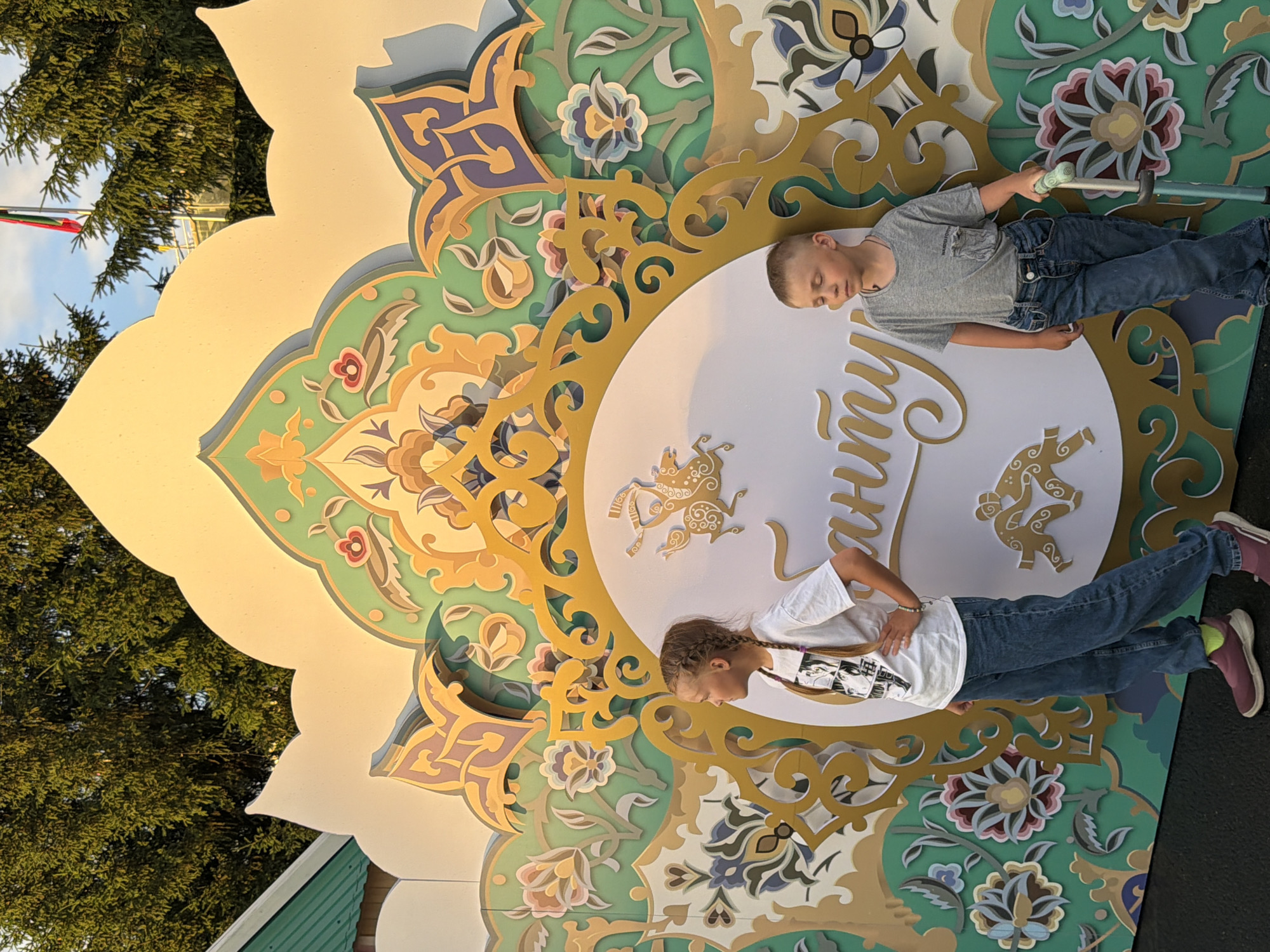 In an outdoor, daytime scene with a golden, ornate backdrop featuring floral and geometric patterns in shades of green, yellow, and blue, two children stand. On the left, a young girl with braided blonde hair, wearing a white t-shirt and blue jeans, stands with her left hand on her hip, looking to her left. On the right, a young boy with short blonde hair, wearing a grey t-shirt and blue jeans, stands with his eyes closed and right hand resting on a teal scooter. Both children are positioned in front of a large circular emblem on the backdrop, which contains decorative gold figures and the word "Санти" in gold cursive. The background includes lush green trees and a glimpse of a flag in the distance. The overall setting suggests a public space or an event.