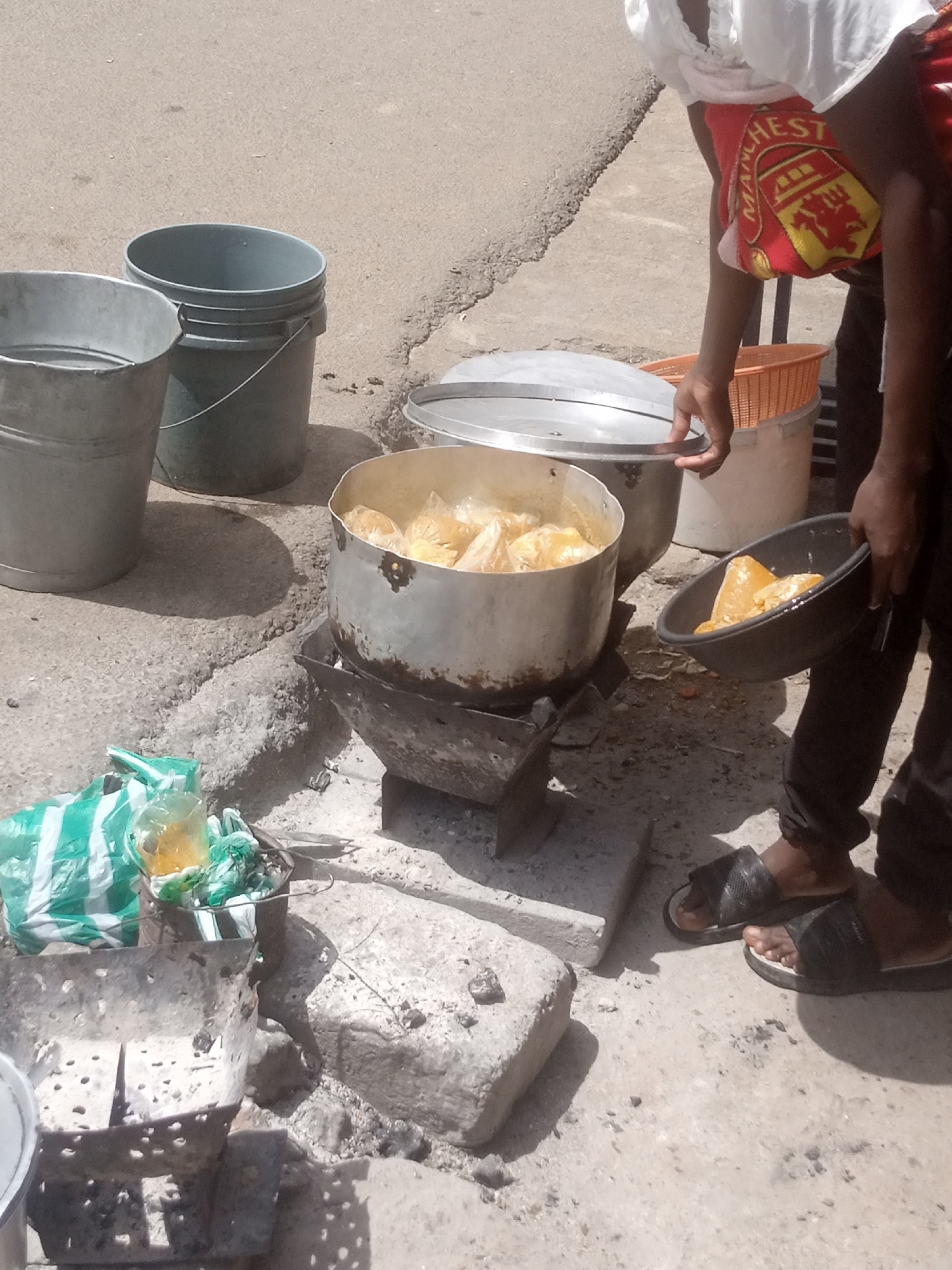The image shows two people outdoors in what appears to be a street setting, possibly in Bukuru, Nigeria. The ground is made of concrete and rough stones. Sunlight casts shadows, indicating it is daytime. The weather appears warm and dry.
One person, wearing dark trousers and black sandals, is holding a dark bowl filled with yellow-orange food items. This person's torso is visible, and they are wearing a white shirt. Around their waist, they have a red cloth with a yellow crest and the text "MANCHESTER UNITED".
Another person's hands are visible, lifting the lid of a large metal pot. This pot is placed on a metal stand, which is itself supported by what looks like concrete blocks. The pot contains more of the yellow-orange food items, some of which are in clear plastic bags.
In the background, there are two large buckets, one silver and one dark grey, and an orange plastic basket. To the left of the large pot, there is a green and white striped plastic bag.
The scene suggests the preparation or serving of food outdoors, possibly for sale or a community gathering. The food appears to be cooked or ready to be served. The setting is simple and practical, with everyday objects like buckets and makeshift cooking stands. There are no strong emotional cues visible, but the activity seems focused and functional.