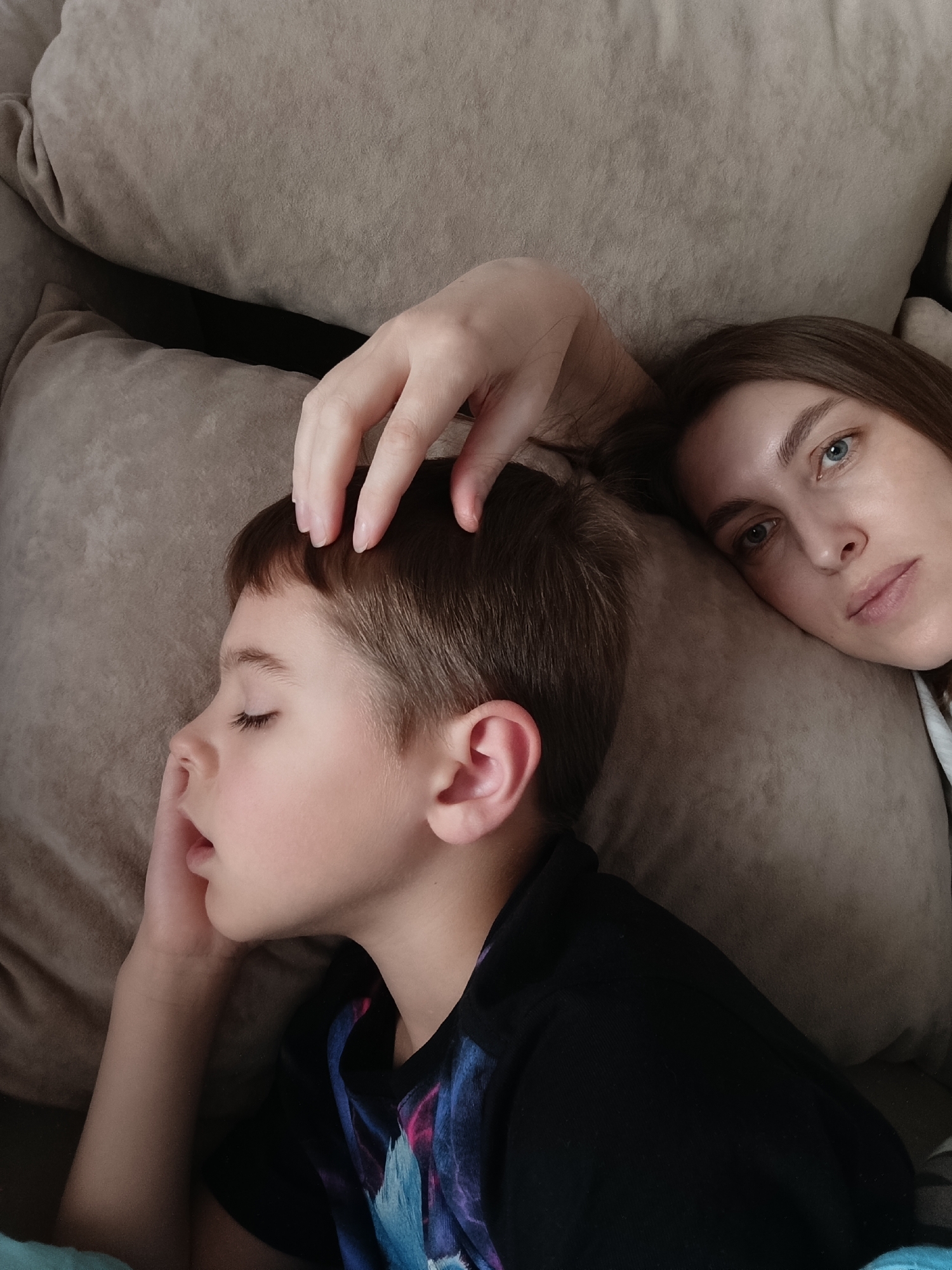 The image depicts a woman and a young boy lying on a light brown sofa. The boy is asleep, lying on his side, with his mouth slightly open and his right hand near his face. He wears a black t-shirt with a colorful design. The woman lies next to him with her head on the sofa, looking directly at the camera. Her right hand rests on the boy's head. The setting suggests a domestic interior. The exact location within the city of Chaikovsky, Russia, cannot be determined from the image.