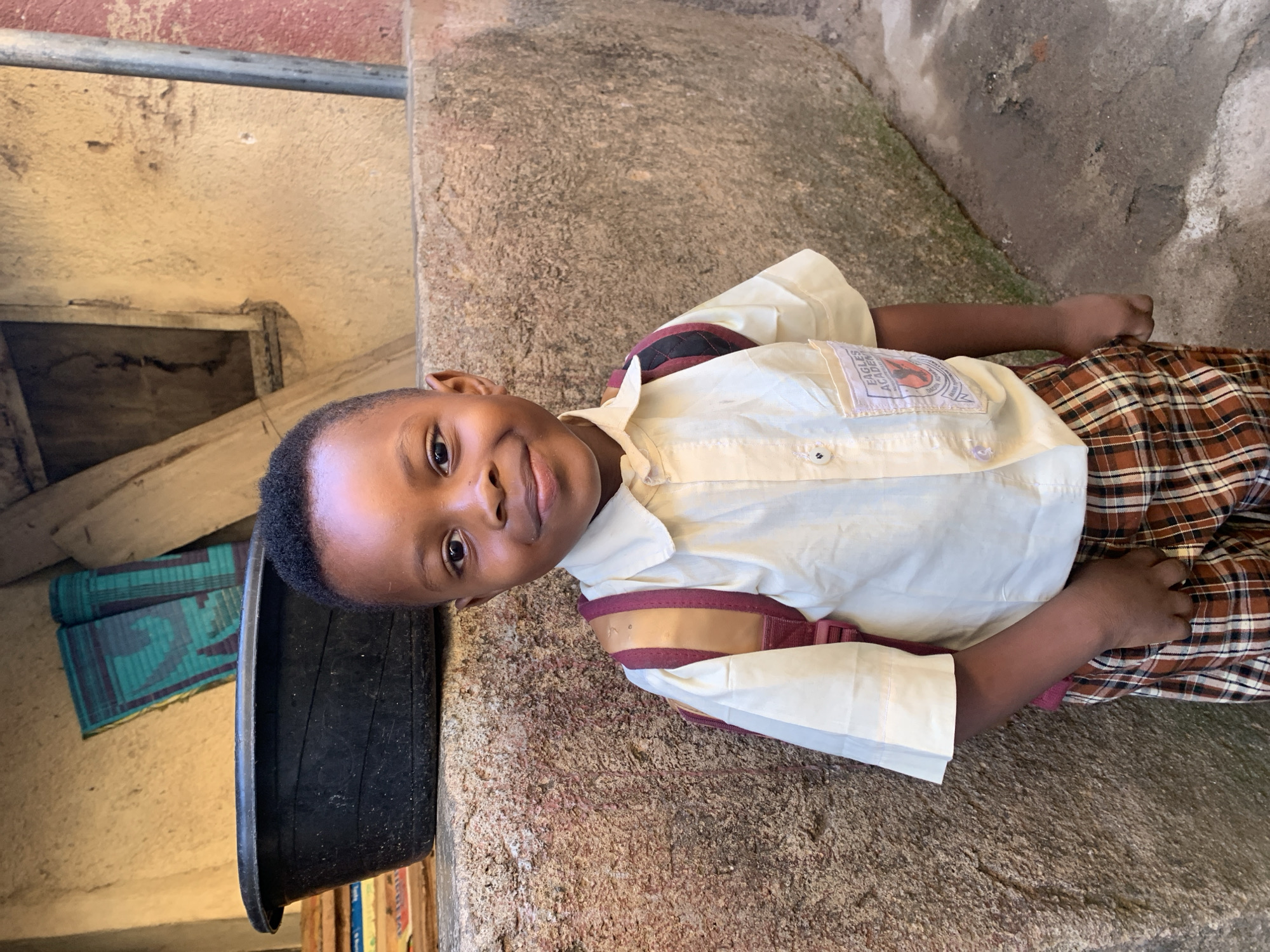 A young boy is lying on a concrete surface. He is wearing a light-colored, short-sleeved shirt with dark trim and plaid pants. A black tub and woven mat are visible nearby. The location is suggested to be in the city of Jos, Nigeria.