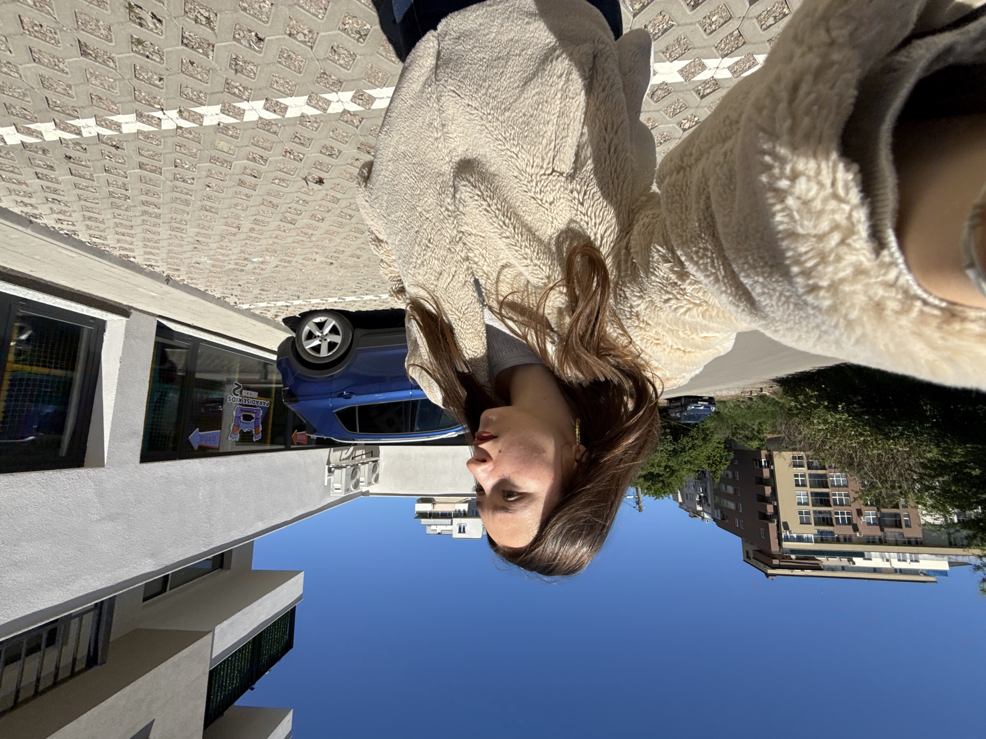 A woman takes a selfie on a sunny day in Sofia, Bulgaria. She is wearing a cream-colored fleece jacket. Behind her, a blue car is parked in front of a building labeled "PARADISE KIDS." Further in the background is an apartment building with trees in front. The sky is clear and blue. A paved area and the facade of a building are also visible.
