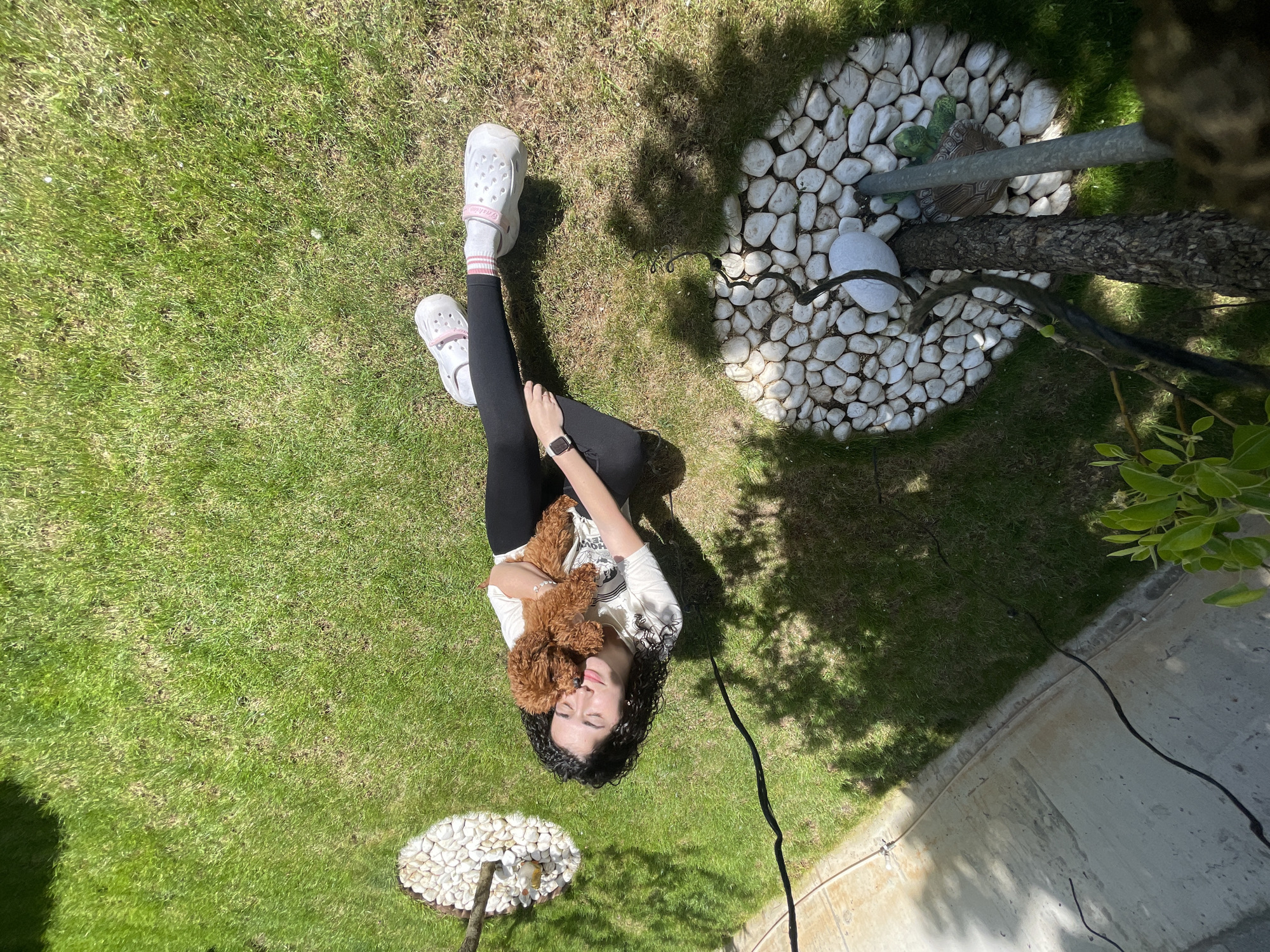 A high-angle outdoor shot shows a young woman lying on green grass, cradling a small, reddish-brown poodle. She is wearing black leggings, a light-colored t-shirt, and white Crocs-style shoes with pink accents. Her dark, curly hair is spread out on the grass behind her head. Her eyes are closed and the dog appears to be resting in her arms.
The location appears to be a backyard or garden. The grass is neatly manicured and interspersed with patches of brown or dry grass. In the background are two circular areas with white stones arranged around what appear to be trees or poles. A thin black wire or cable runs from the lower right corner of the frame, across the grass, towards the center. The lighting suggests a sunny day, with shadows visible on the grass. Based on the architectural elements in the background, the setting is likely in a residential area within the city of Sofia, Bulgaria.