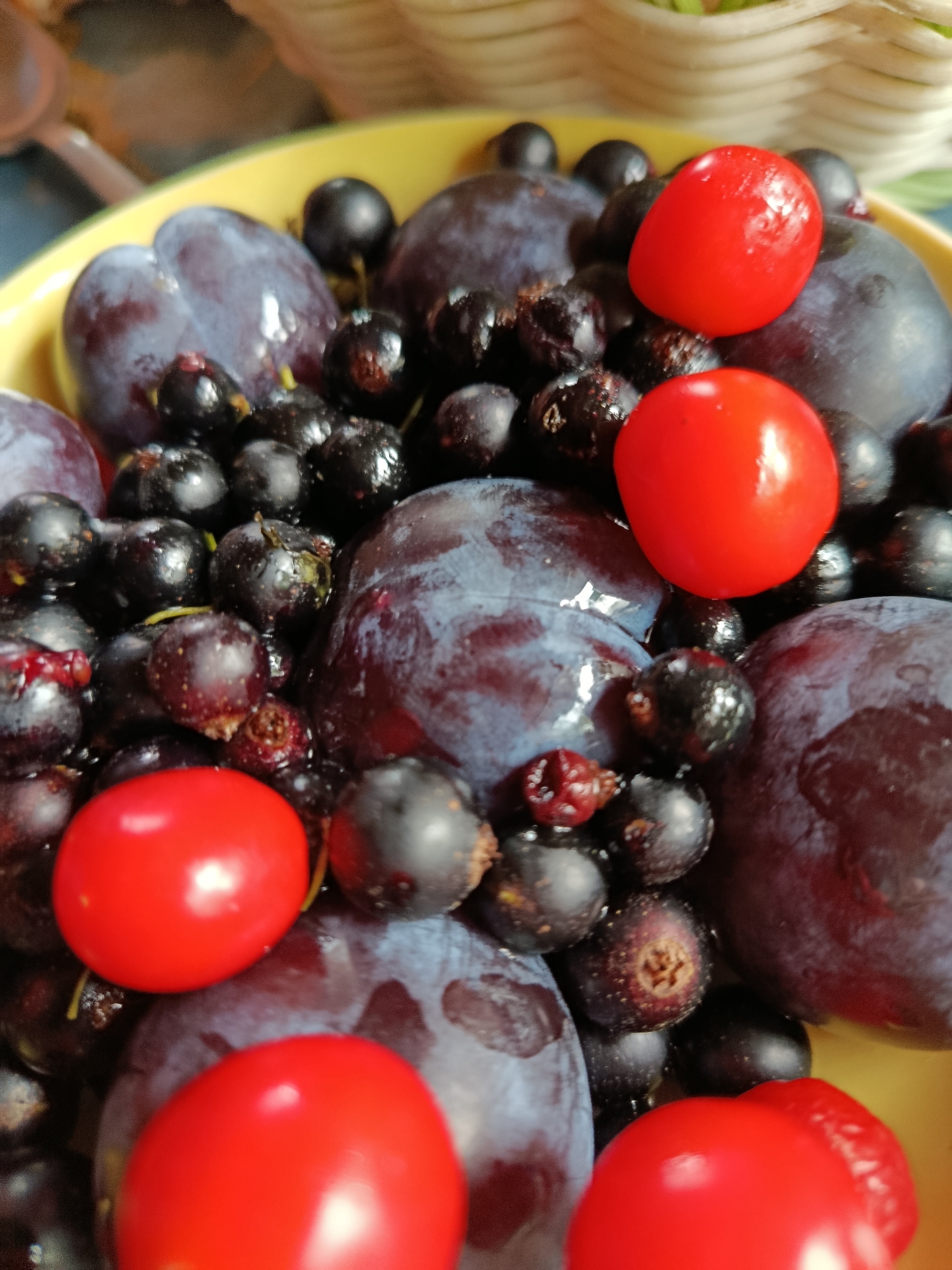 A close-up, high-angle shot shows a pile of fruits in a yellow bowl. The fruits include several dark purple plums, numerous small black currants, and a few bright red cherry tomatoes. The fruits are densely packed, with some plums partially obscuring the currants and tomatoes. The lighting is somewhat diffused, creating soft highlights on the fruits. The background is out of focus, with hints of a woven basket and a blue surface. There is no indication of a specific location in the city of Верея, Russia.