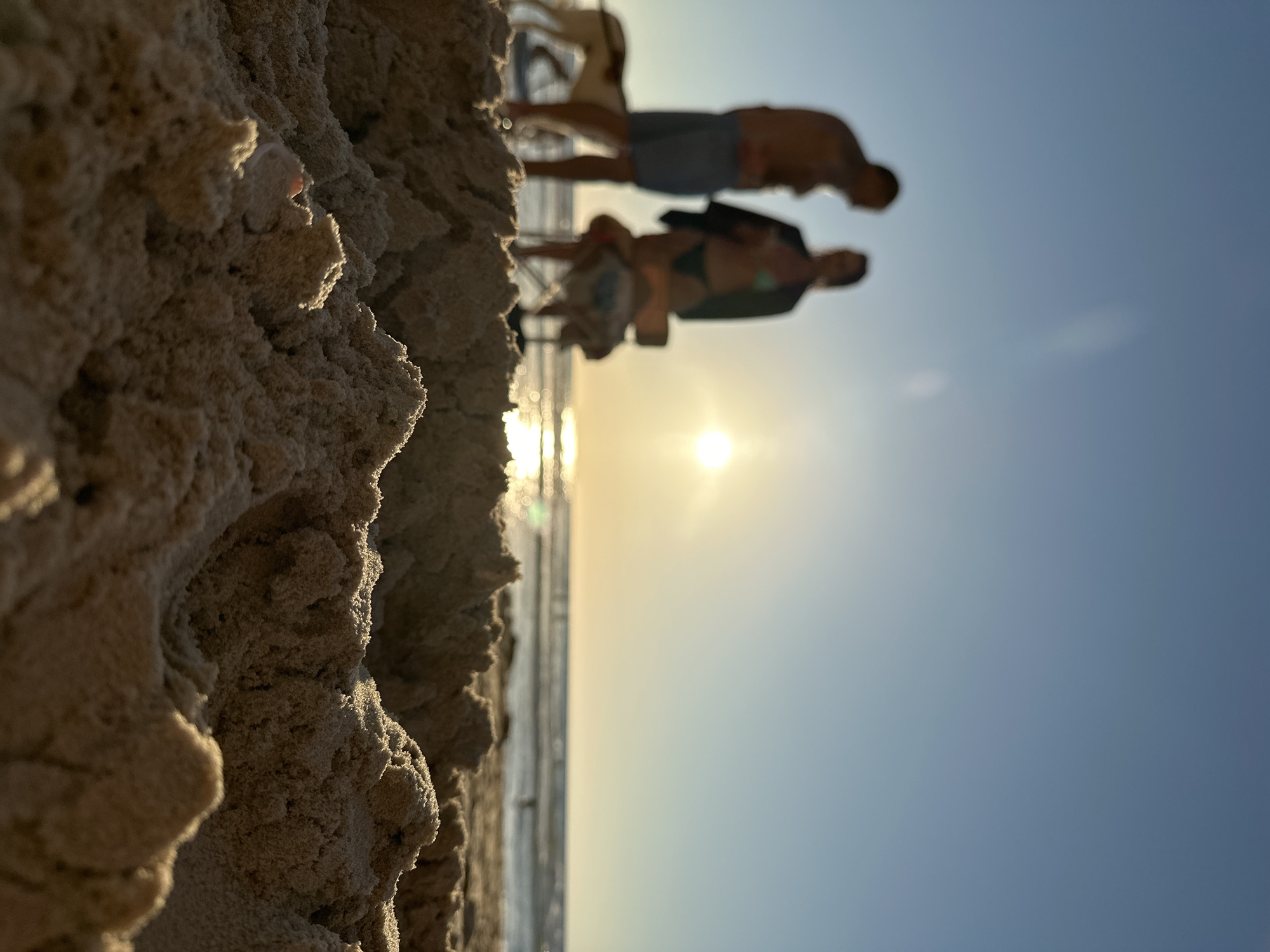 The image depicts a sandy beach in Herzliya, Israel, at sunset. The foreground is dominated by textured sand dunes, obscuring the lower portion of the scene. In the mid-ground, the sun is setting over the ocean, casting a warm glow. Silhouetted against the bright sun are two people and a dog. One person, likely male, stands on the left wearing shorts. The other person, likely female, stands to the right, wearing a bikini and a dark cover-up. Between them, a beach chair is visible. The ocean stretches to the horizon, with faint figures of people in the water. The sky is a clear gradient of blue, transitioning to a lighter hue near the horizon where the sun is setting.