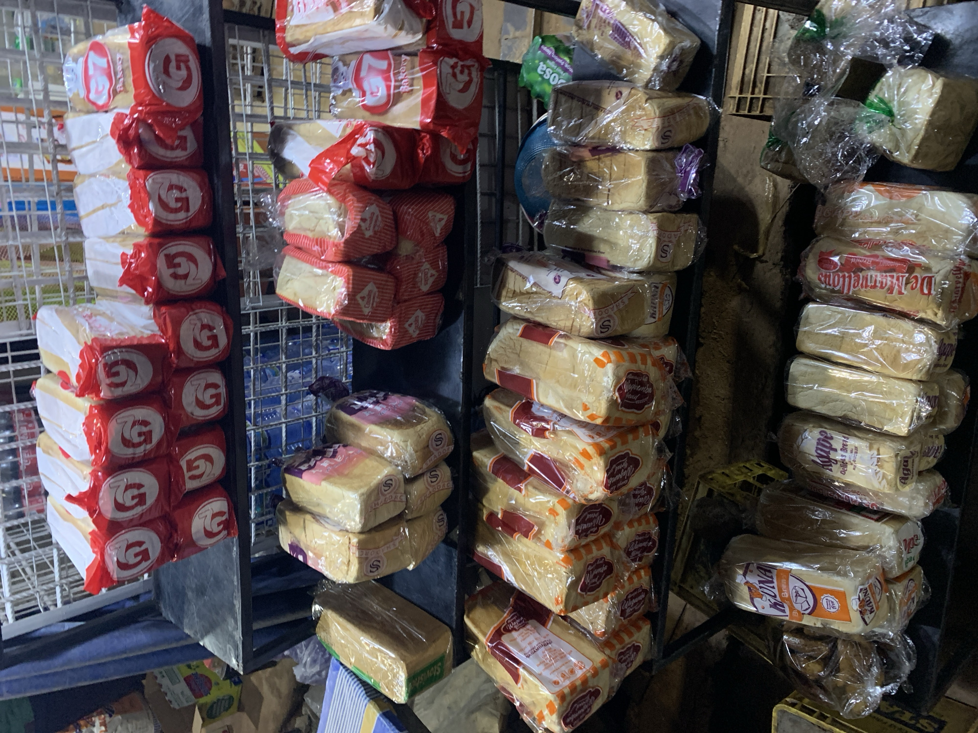 The image depicts a retail display of various loaves of bread, predominantly in clear and red-and-white wrappers, hanging from metal racks. The bread loaves are stacked vertically, with different brands and packaging visible. Some of the brands visible include "G7 Bakery," "Shadorc," "Delicas Bread," and "Agape." The location is identified as the New Stadium area in Nigeria. The scene appears to be a roadside or market stall due to the dark and uneven lighting, along with the presence of crates and miscellaneous items visible in the background. No people are visible in the frame.