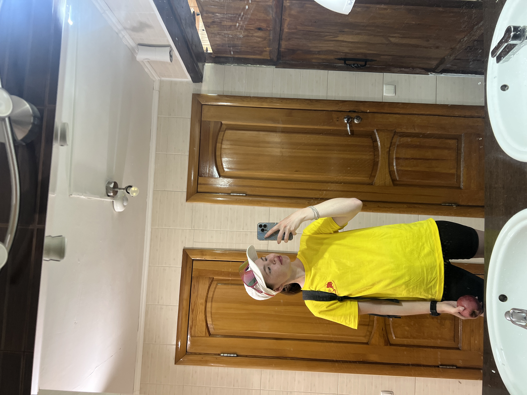 A young person is taking a selfie in a bathroom. They are wearing a yellow t-shirt, black pants, and a baseball cap. They are holding a red object, possibly an apple, in their left hand. The bathroom has a white sink, a wooden cabinet, a wooden door, and tiled walls. The image was likely taken in Khvalynsk, Russia.