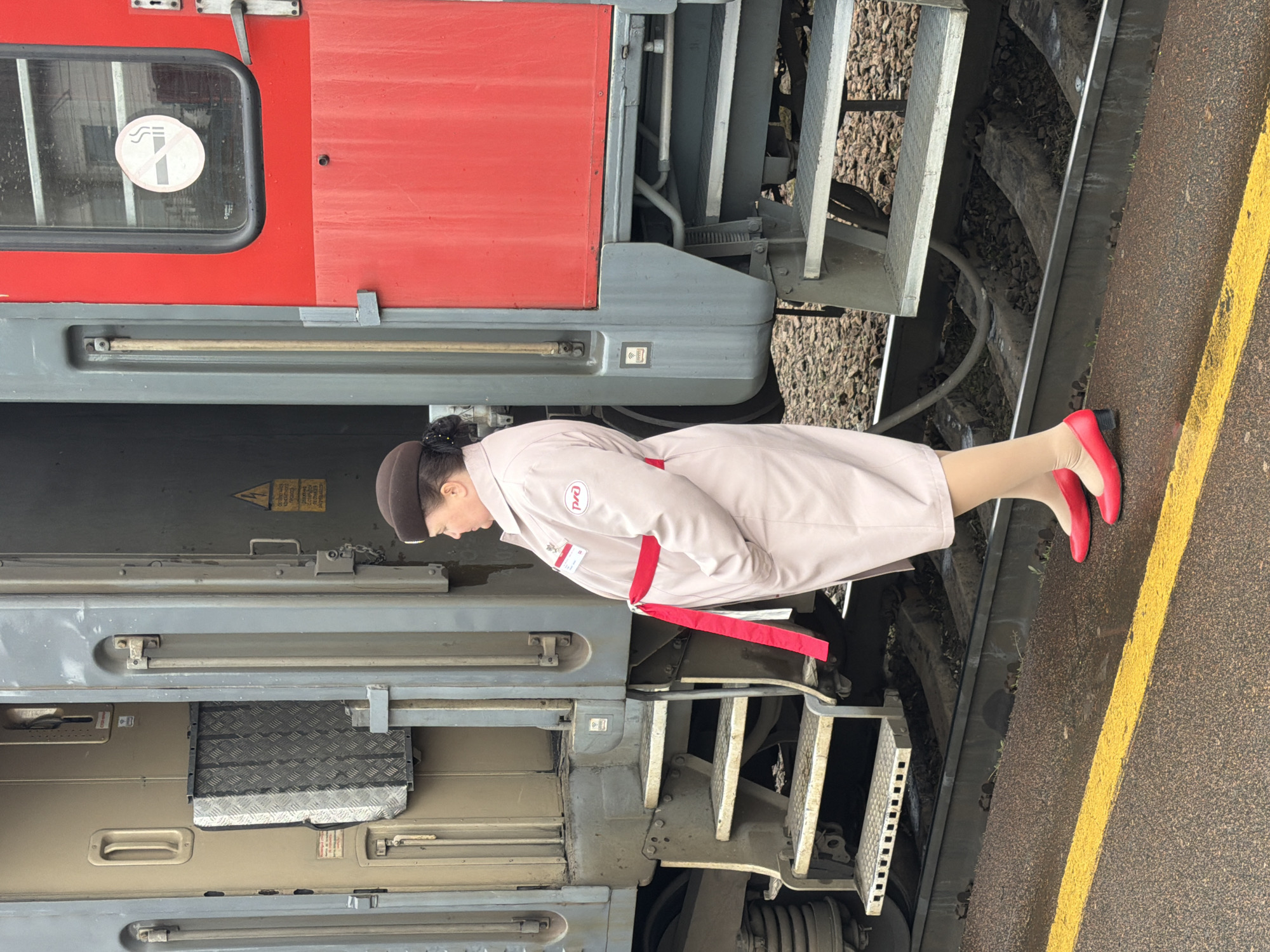 At a train station in Liski, Russia, a woman wearing a light tan uniform with red accents, a brown hat, and red heels stands beside a red and gray train car. The train is on the tracks, with a yellow safety line visible on the platform. The woman is facing towards the train and looking down. The train car has metal steps and a non-smoking sign in the window.