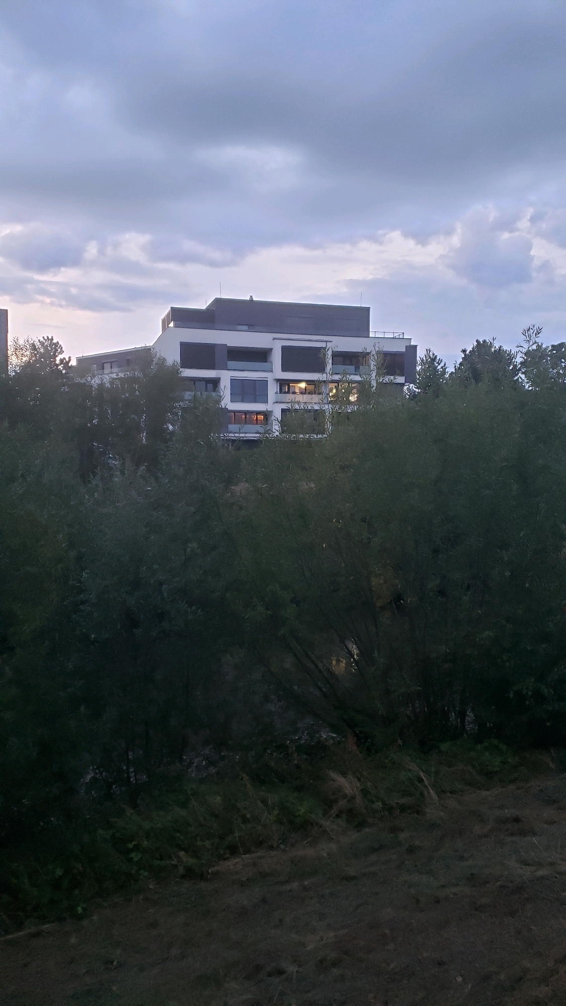 The image shows a modern, multi-story building with a white facade and dark gray accents, partially obscured by dense green foliage. The building has multiple windows, some of which are illuminated, suggesting occupancy. Above the building, the sky is overcast with gray clouds. The foreground consists of dark, lush vegetation and a patch of dry grass. The scene appears to be a residential area, possibly on the outskirts of the city of Prešov, Slovakia, given the architectural style and the surrounding natural elements. There are no people visible in the image.