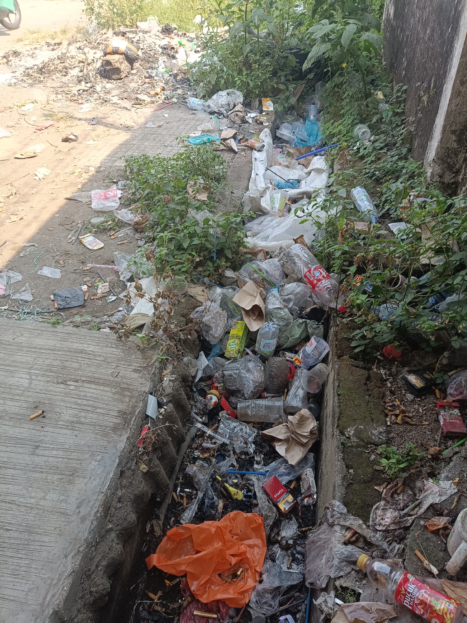 The image depicts a roadside drain overflowing with a significant amount of mixed waste in Jember, Indonesia. The drain, situated alongside a paved area and a concrete structure, is choked with plastic bottles, plastic bags, food wrappers, cigarette butts, and other miscellaneous debris. Overgrown vegetation is present, intertwining with the garbage. The scene suggests a lack of proper waste management, leading to environmental pollution in a semi-urban setting.