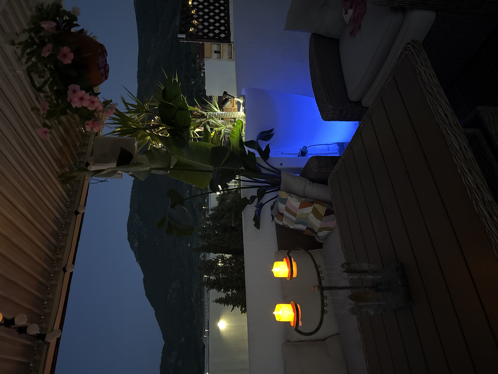 The image depicts an outdoor patio area at dusk, with a mountainous landscape in the background. The patio is furnished with a sectional sofa with white cushions, a wicker armchair, and a wooden table. Two LED candles on a small side table emit a warm, orange glow. Lush green plants in a large, illuminated planter with blue light are also visible. Decorative string lights hang overhead. The scene suggests a tranquil evening setting in Vallada, Spain. There are no people visible in the image.