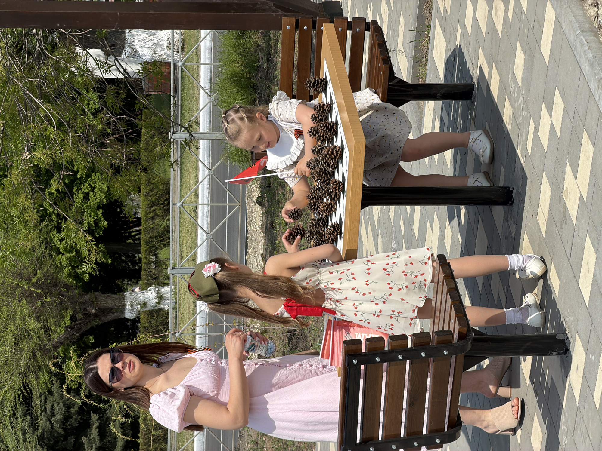 The image shows a young woman and two young girls in an outdoor setting. The woman is standing on the left, wearing a light pink dress and sunglasses. She is holding a water bottle. One of the girls is sitting on a bench, wearing a dress with a red floral pattern. She is also wearing an olive green cap with a pink flower. The second girl is sitting on the other side of the bench, wearing a light dress with a white collar and a red ribbon. They are playing a game on a chess/checker board that is filled with pinecones. There is a red flag with a floral design stuck into the board. The location appears to be a paved area in Kamyshin, Russia. There is greenery and fencing in the background.