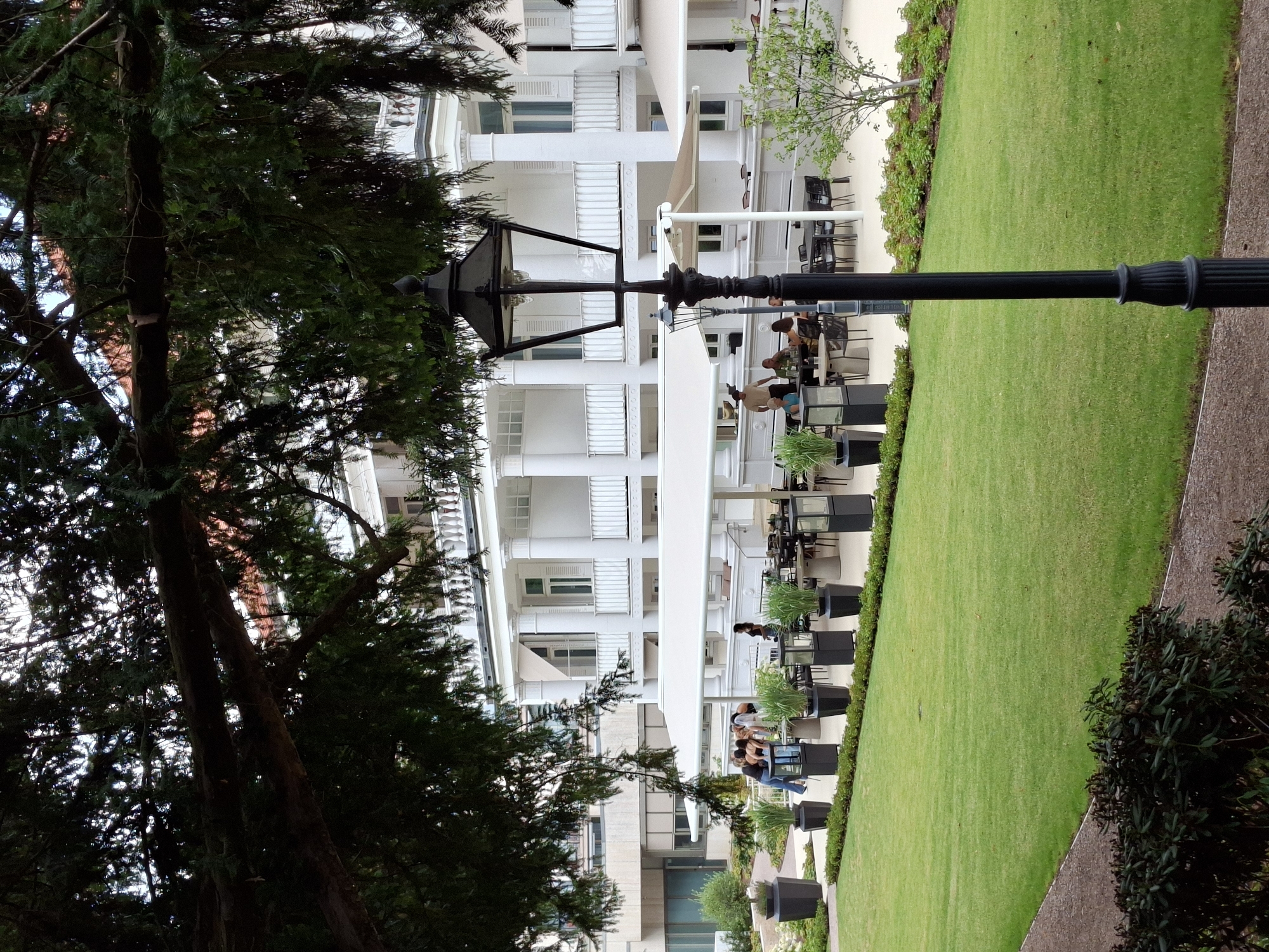 A group of people are gathered at tables outside a white building with columns and balconies. The building appears to be a hotel or a restaurant. A street lamp stands in the foreground, and a lawn is visible in the background. The scene is set in Baden-Baden, Germany.