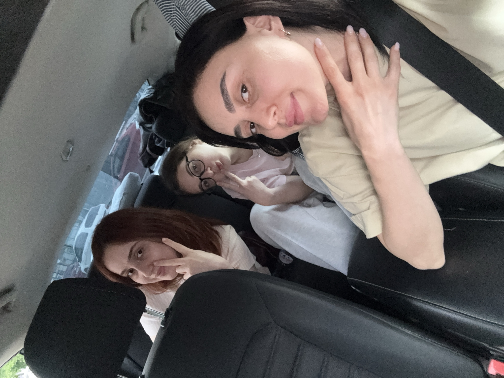 Three people are in the back seat of a car. The car is likely in the city of Tbilisi, Georgia, as suggested by external features. The person in the front, with dark hair and a seatbelt, is looking at the camera and holding up her hand. Behind her, another person wearing glasses is doing the same. In the background, a third person with red hair is posing with a "peace" sign.