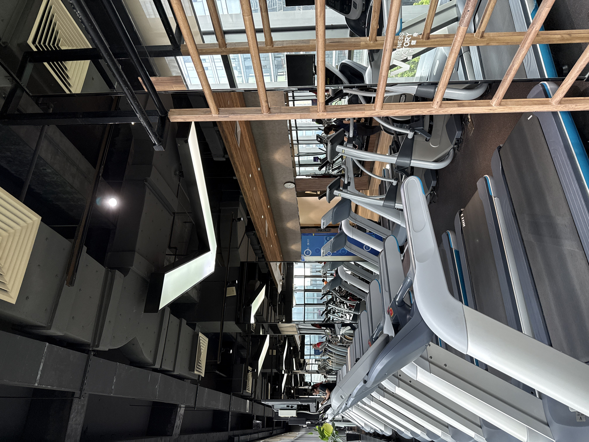 This image depicts the interior of a modern gym located in Haizhu District, China. The scene is dominated by rows of treadmills, with their grey and white frames and black running belts filling the foreground and mid-ground. Several people are visible in the background, appearing to use or walk past the exercise equipment. The gym features an industrial-style ceiling with exposed black ducts and ventilation systems, contrasted by sleek, modern light fixtures. Mirrors line one wall, reflecting the gym equipment and further enhancing the sense of space. To the right, a wooden climbing frame with horizontal bars stands against a mirrored wall, behind which can be seen large windows offering a view of urban buildings. The overall environment suggests a well-equipped and contemporary fitness facility.